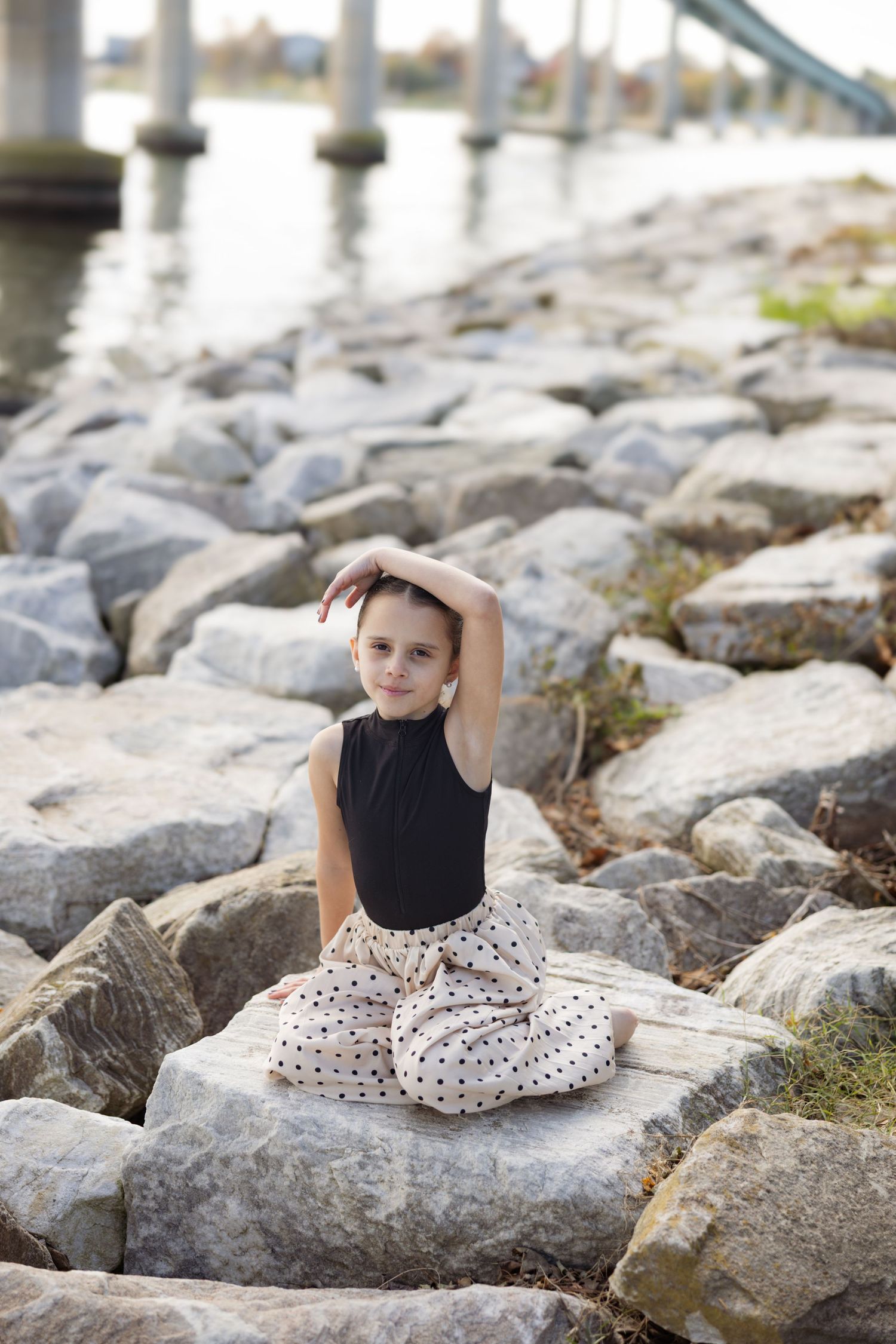 A person in a black top and patterned skirt sits gracefully on large coastal rocks near the water.
