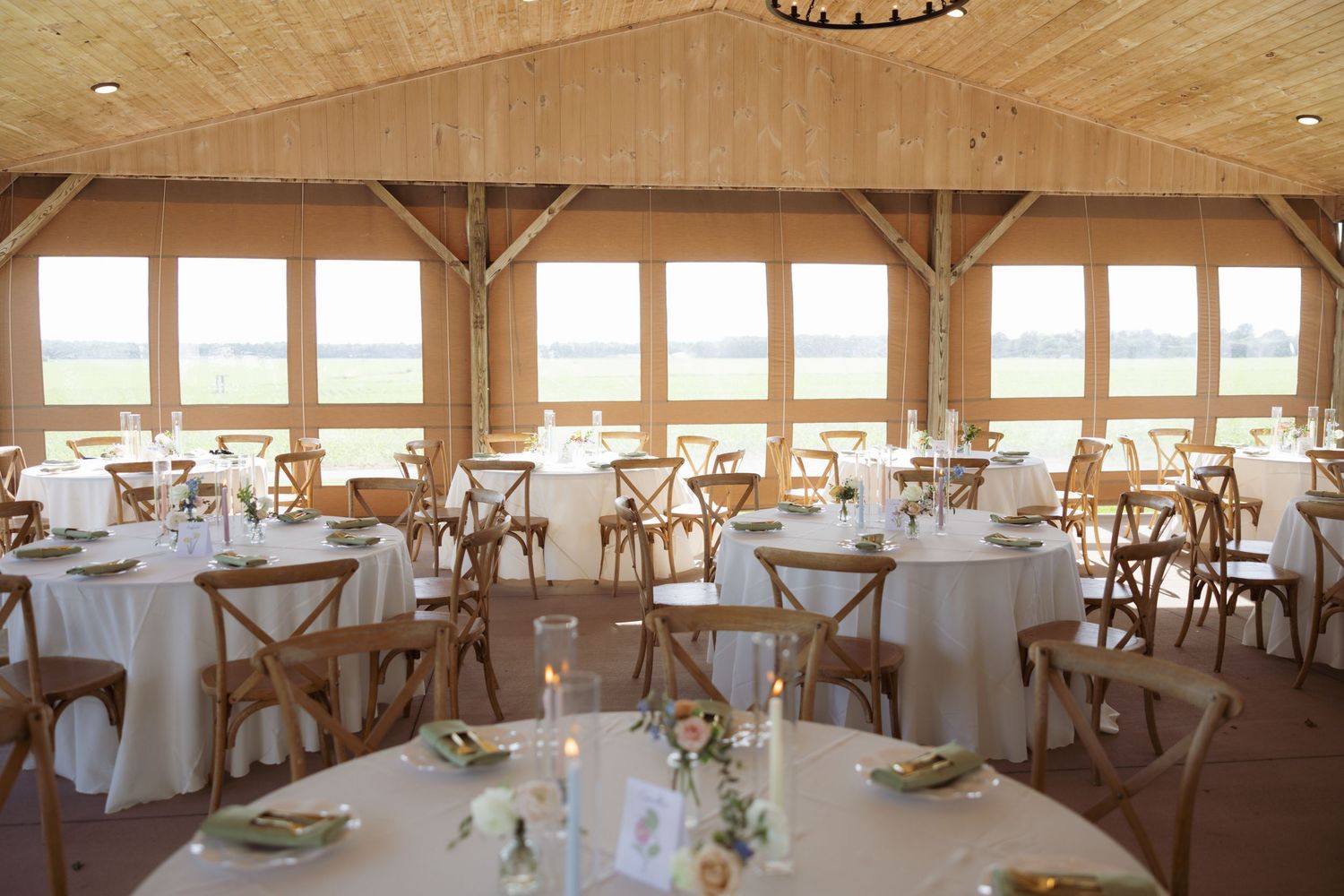 Elegant barn wedding reception hall with round tables, white linens, wooden chairs, and panoramic windows overlooking a scenic waterfront.