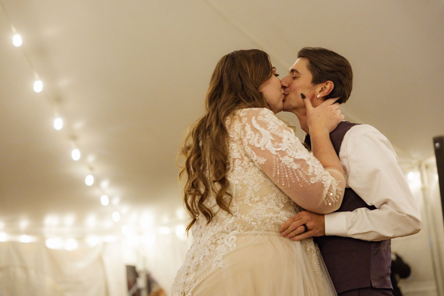 A bride and groom share a romantic first kiss at their wedding reception, surrounded by warm string lights.