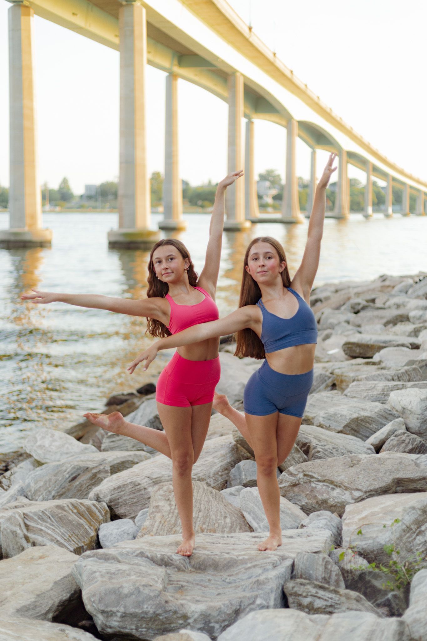 Two dancers strike graceful poses on rocky riverbank beneath a bridge at sunset.