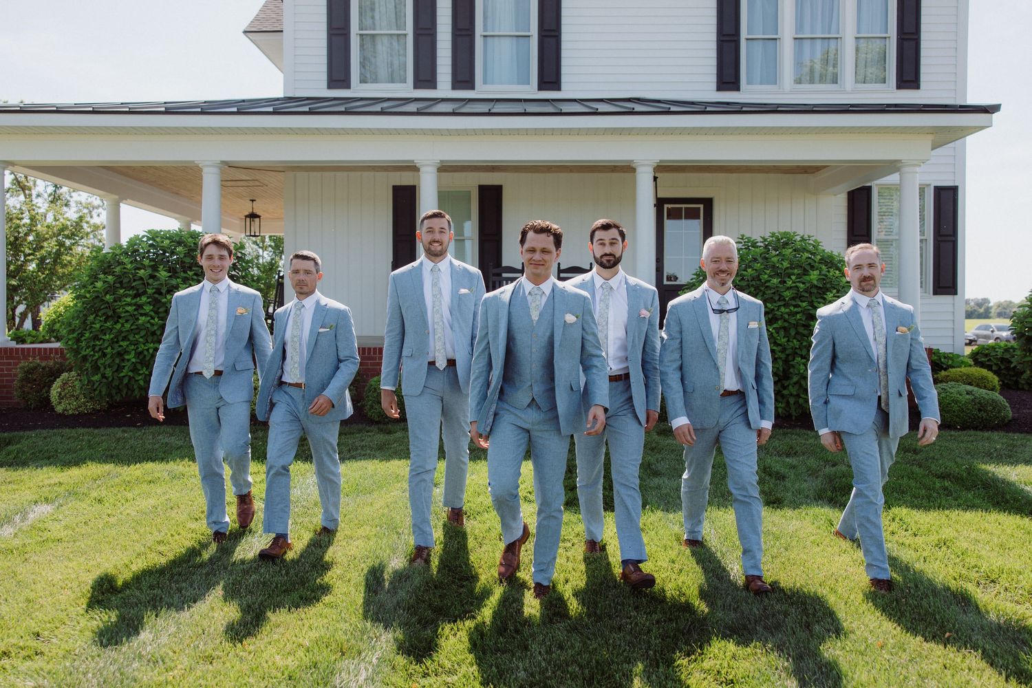 Groomsmen in light blue suits walk together outdoors in front of a white farmhouse on a sunny wedding day.
