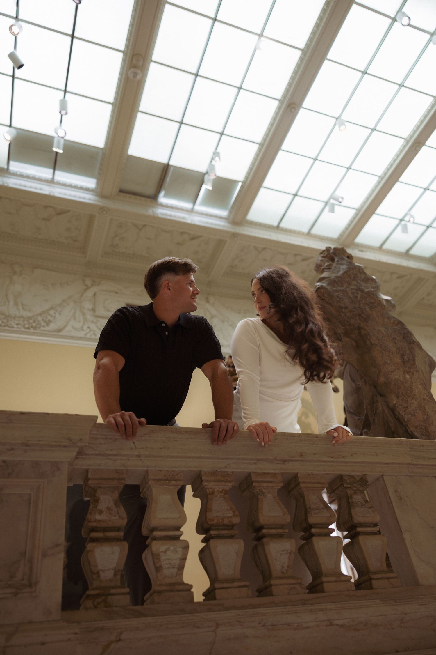 Two people lean over an ornate marble balustrade in a light-filled interior space with decorative architectural details.