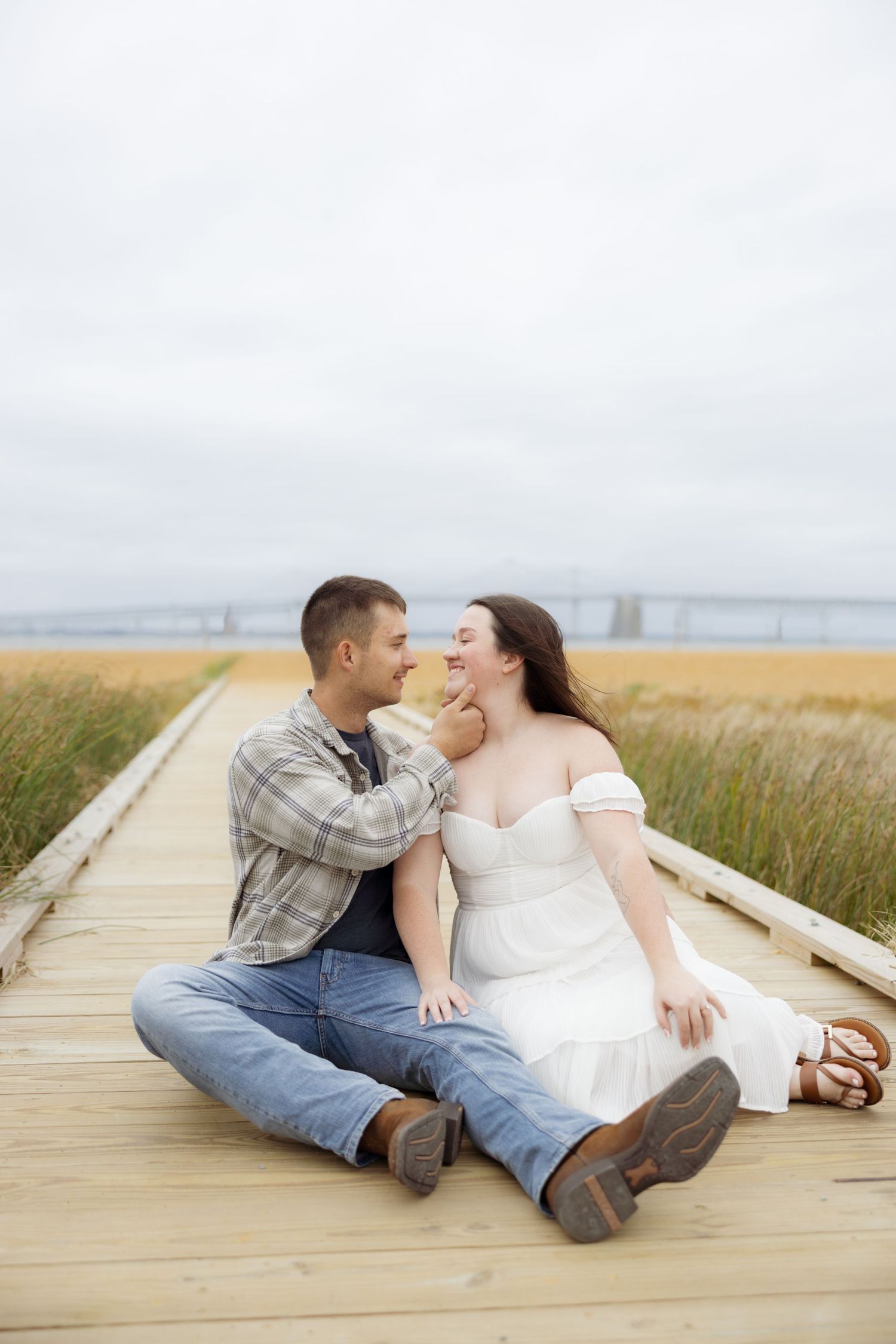 A couple shares romantic moments while sitting on a wooden boardwalk through a grassy coastal area.
