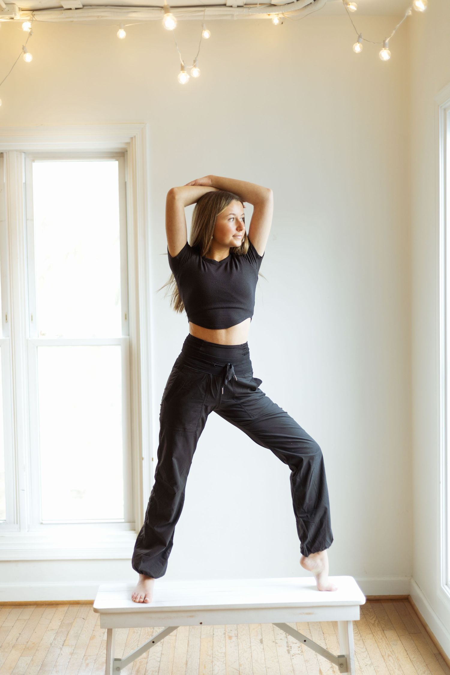 Dancer in black outfit stretches gracefully on white bench against bright window and string lights.