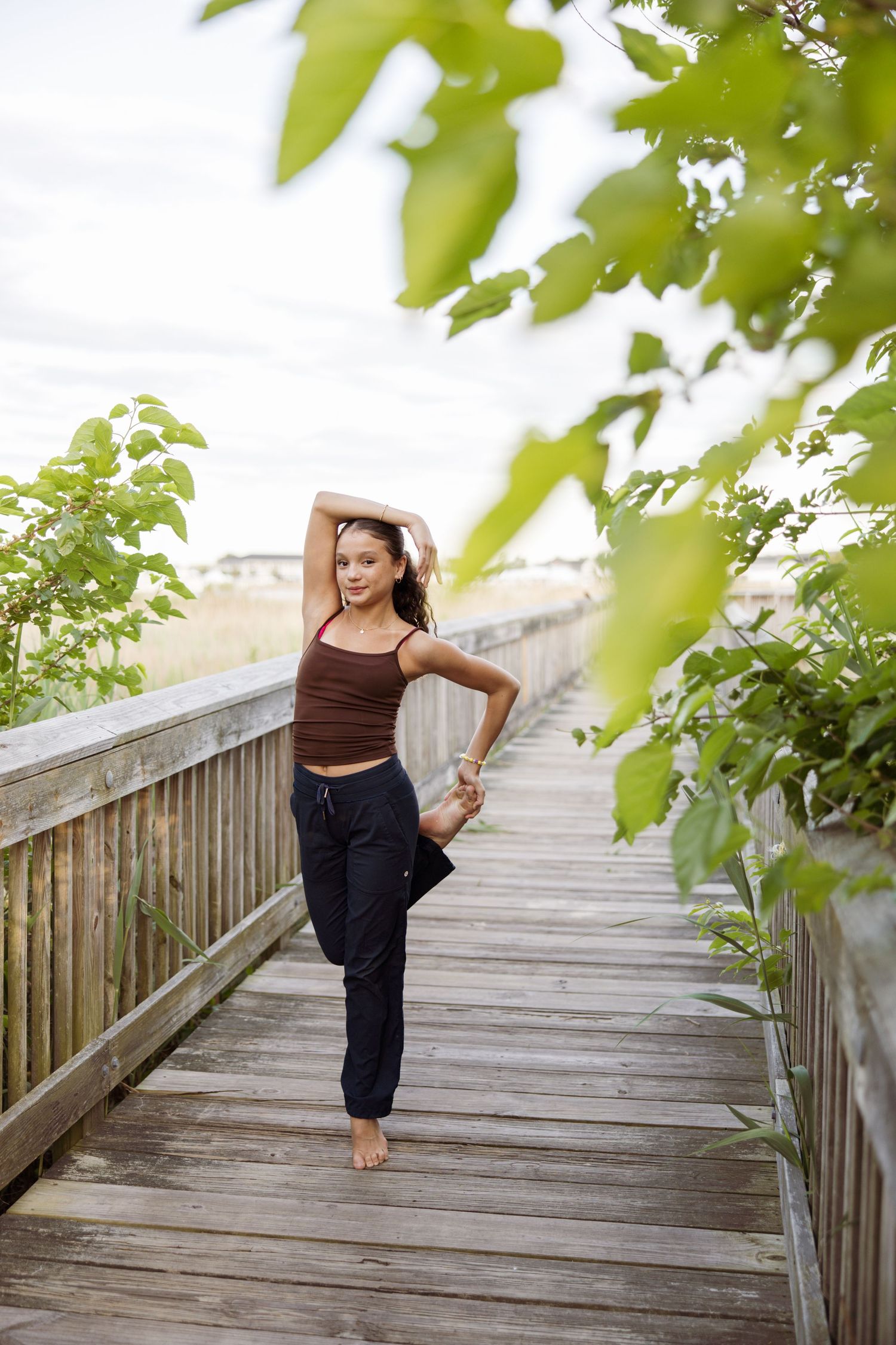 Young dancer practices ballet pose on wooden boardwalk surrounded by green summer foliage.
