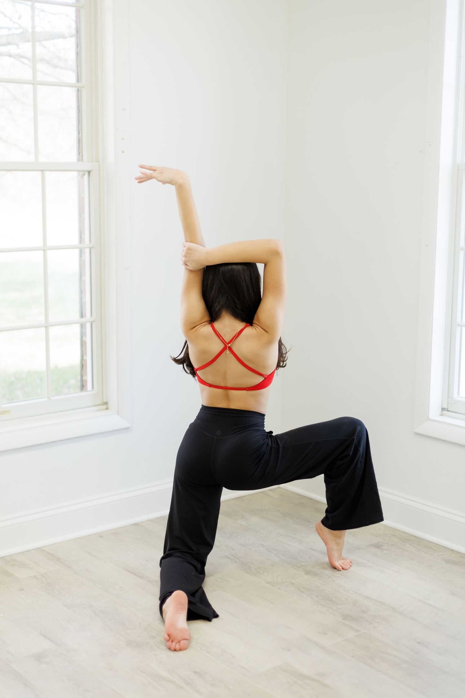 Dancer strikes elegant pose with raised arms while wearing red crossback top in minimalist studio setting.