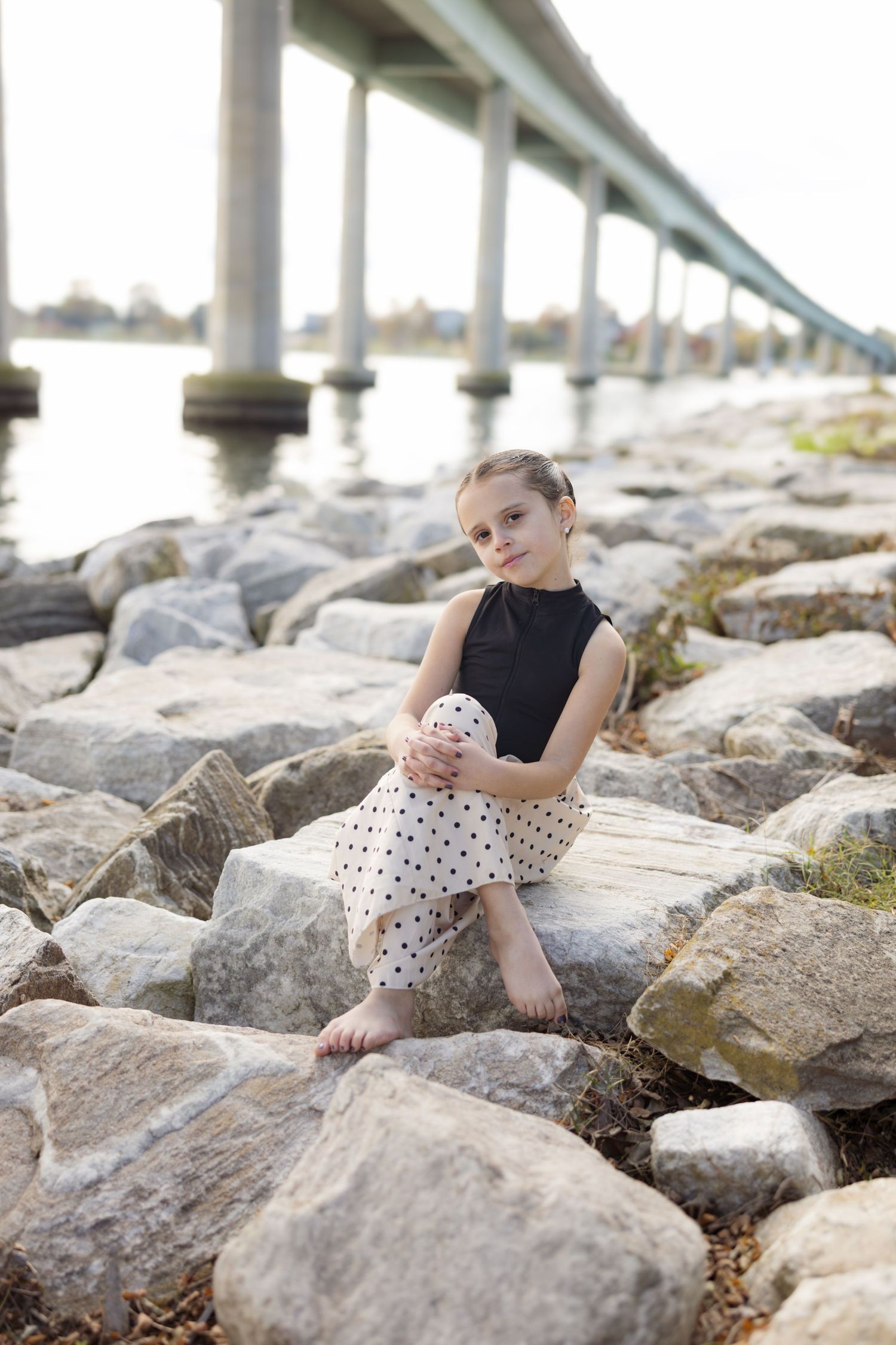 A black sleeveless top and polka dot skirt outfit photographed on rocky shoreline beneath a bridge.