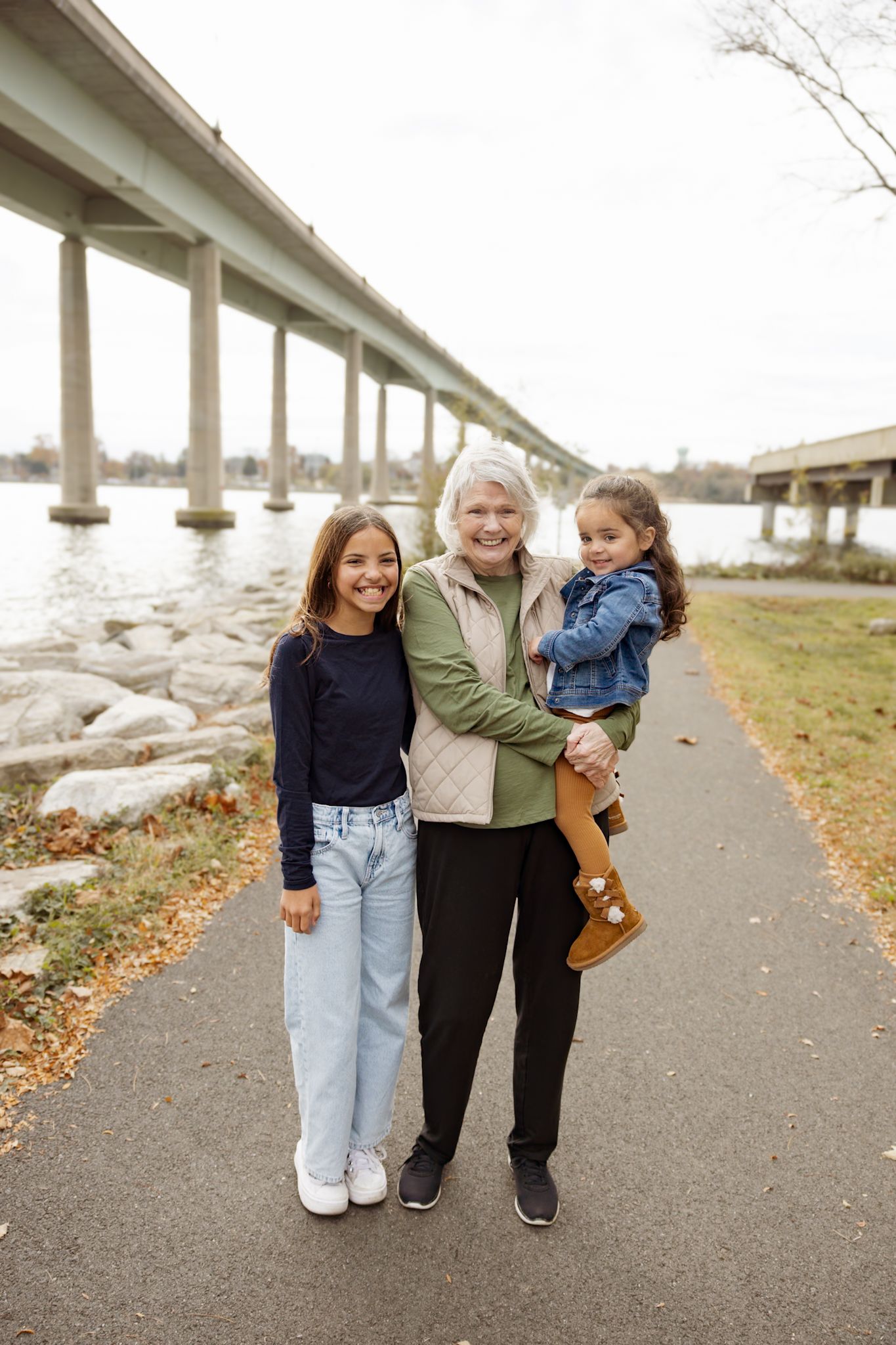 Three people standing on a path by a bridge over water on an overcast day.