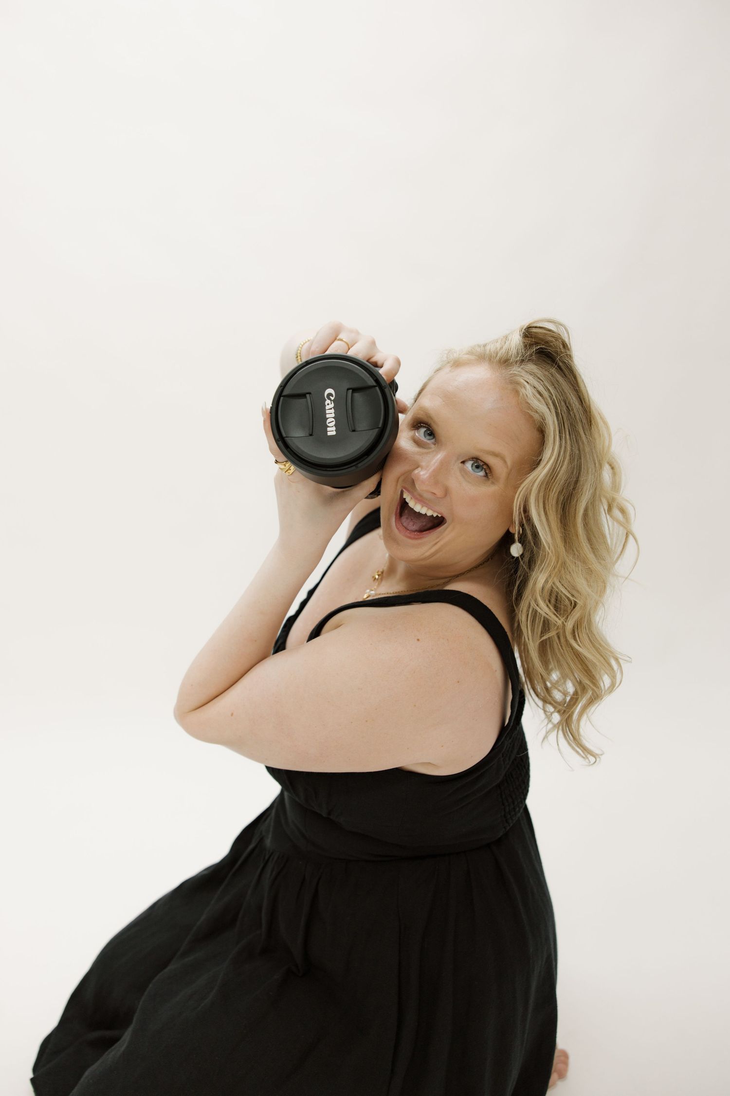 Photographer with wavy blonde hair holds camera lens while posing joyfully in black dress against white backdrop.