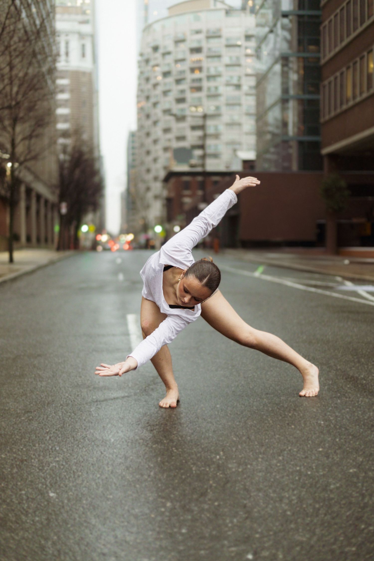 Modern dancer executes contemporary move on rainy city street between tall buildings.