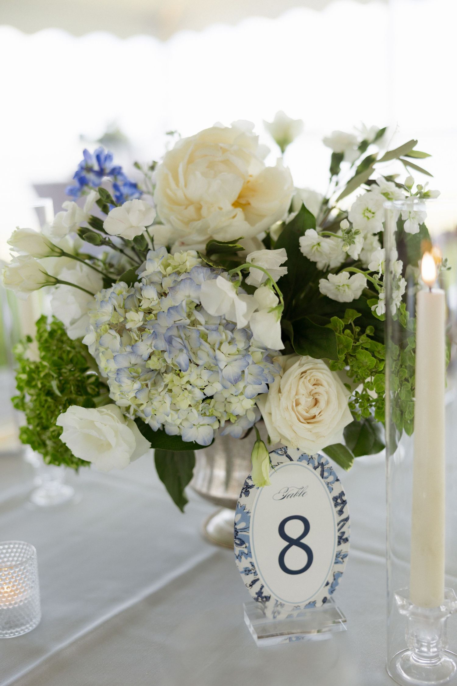 Elegant wedding centerpiece with white peonies, blue hydrangeas, and roses in silver vase beside table number 8 card and candles.