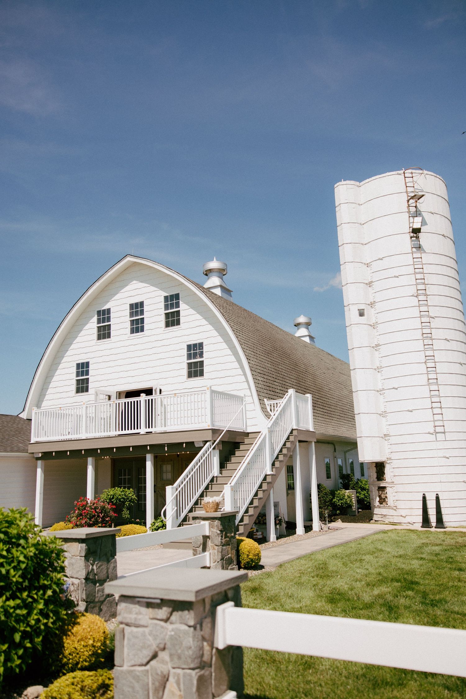 White barn wedding venue with gambrel roof, silo, wraparound deck, and manicured grounds under blue sky.