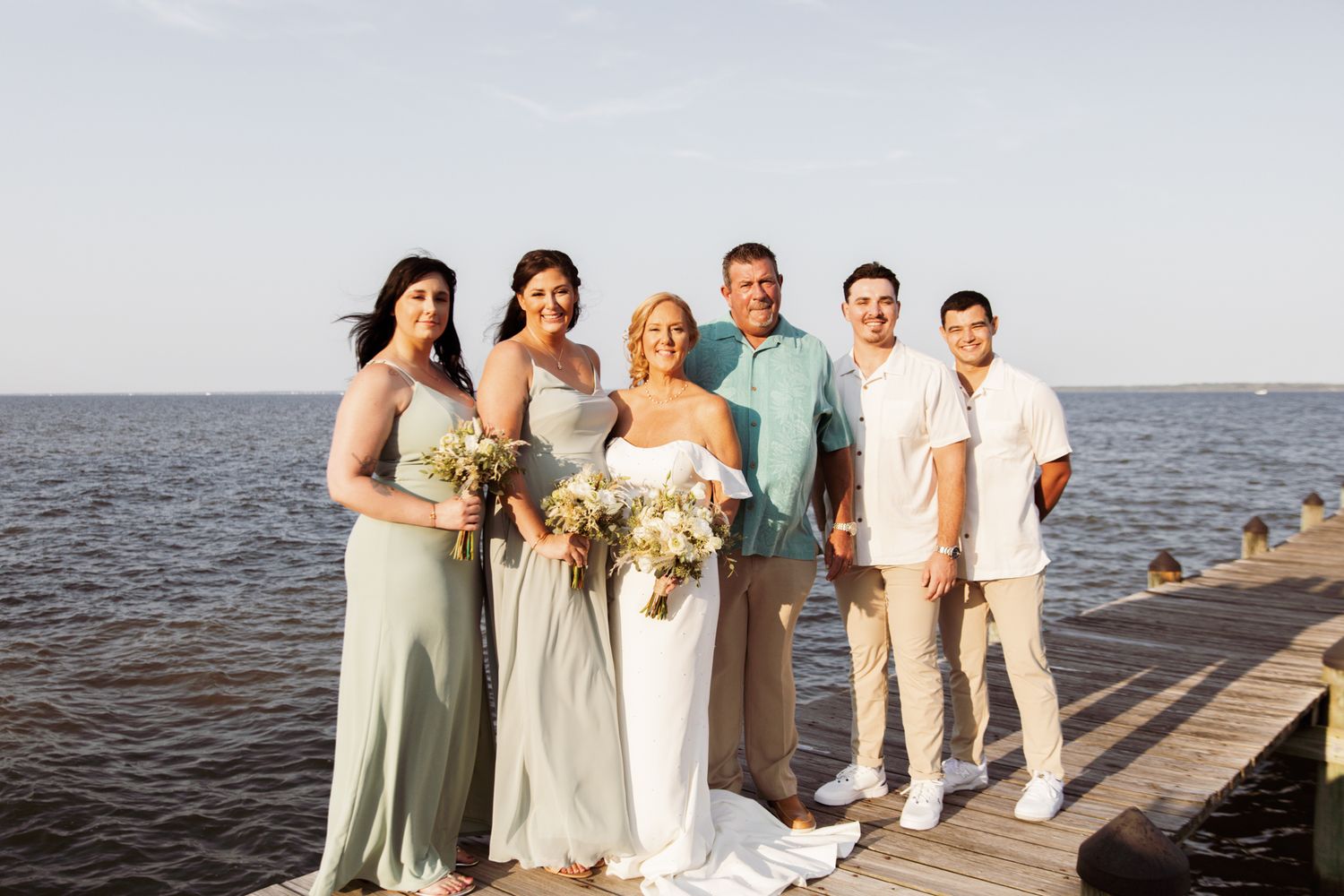 Wedding party posing on a waterfront dock, featuring bridesmaids in sage green gowns and a bride in white holding bouquets.