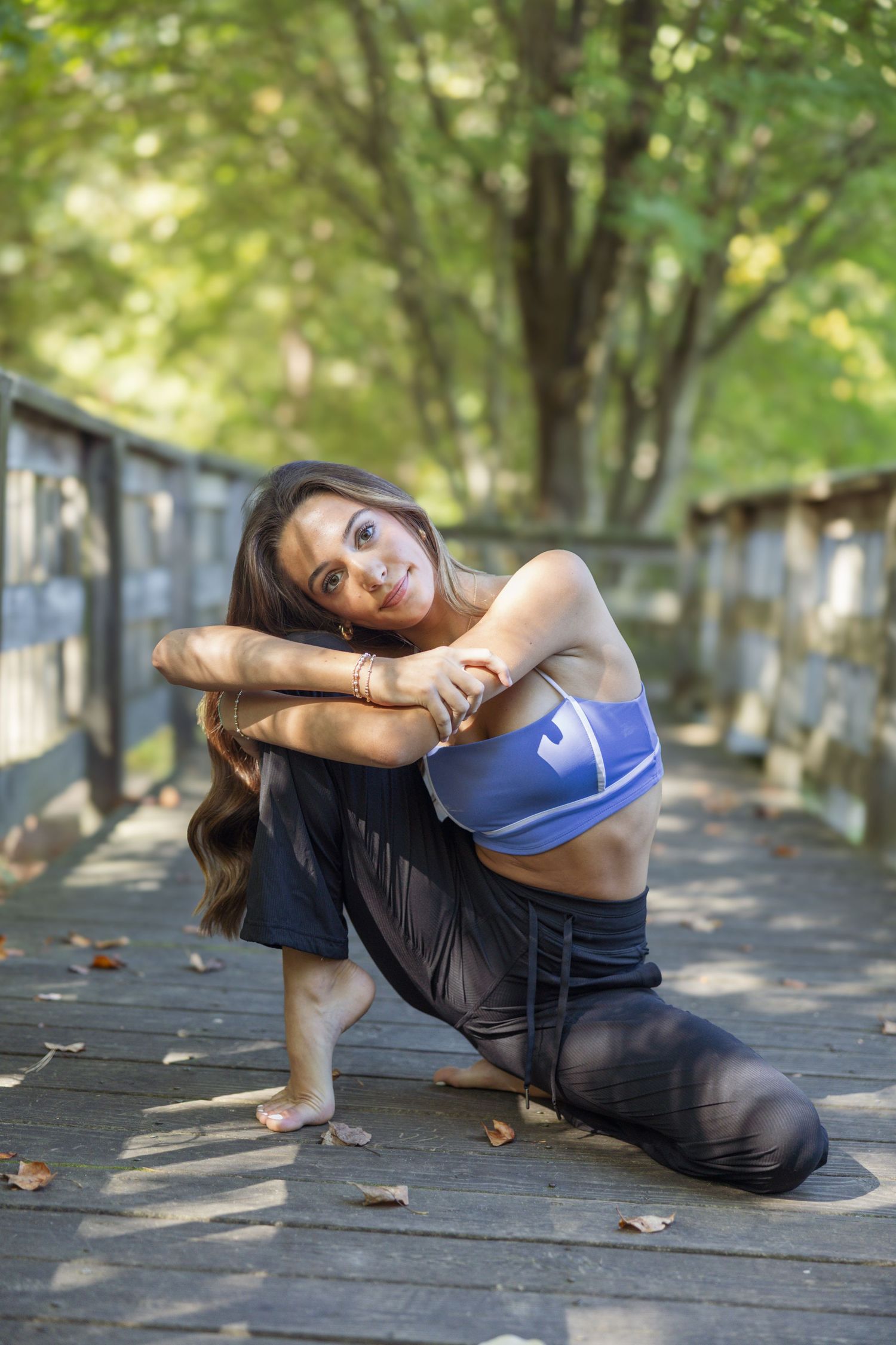 A dancer in athletic wear strikes a graceful pose on a wooden bridge surrounded by lush green trees.
