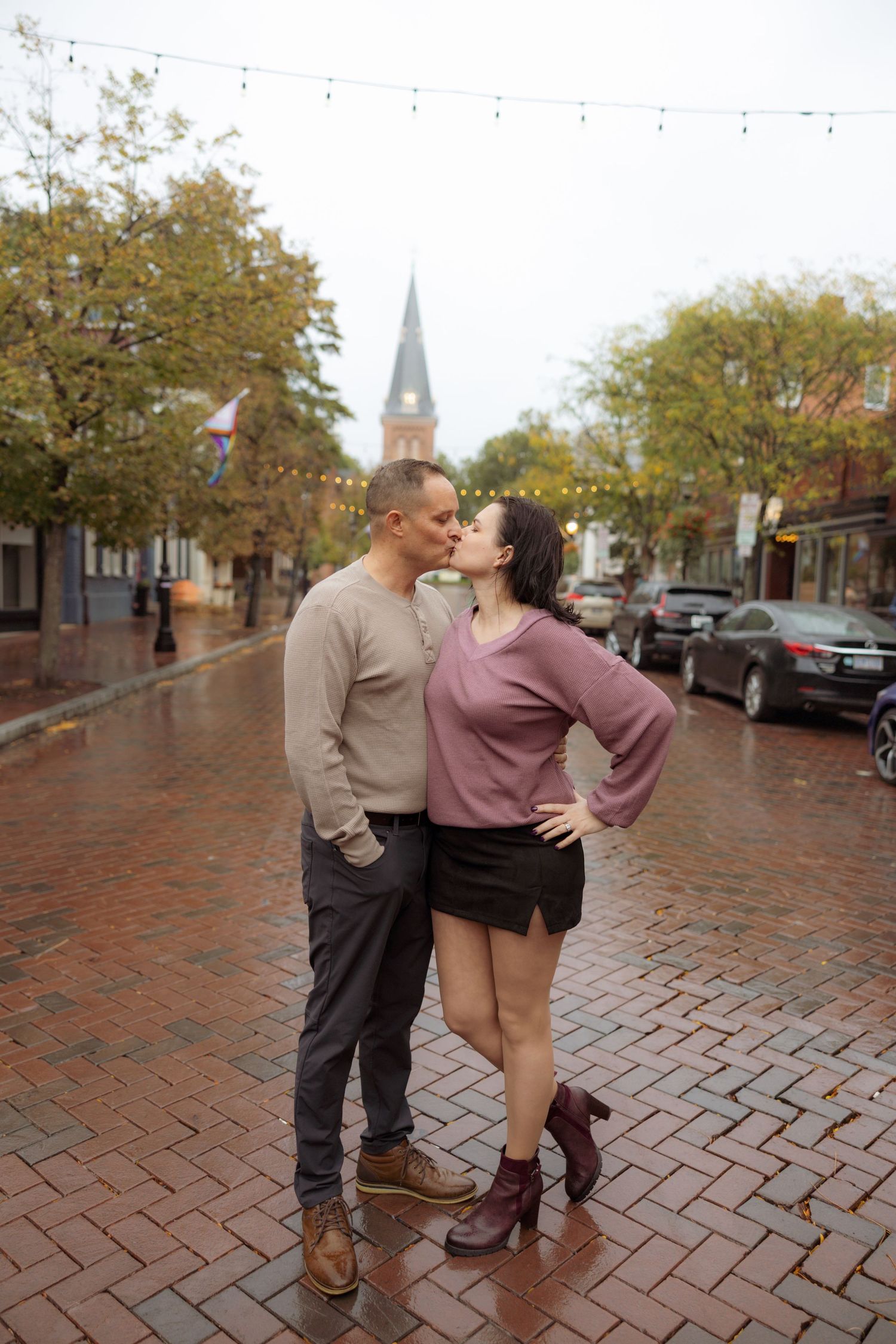 A couple shares a romantic moment on a brick-paved street with autumn trees and a church spire in the background.
