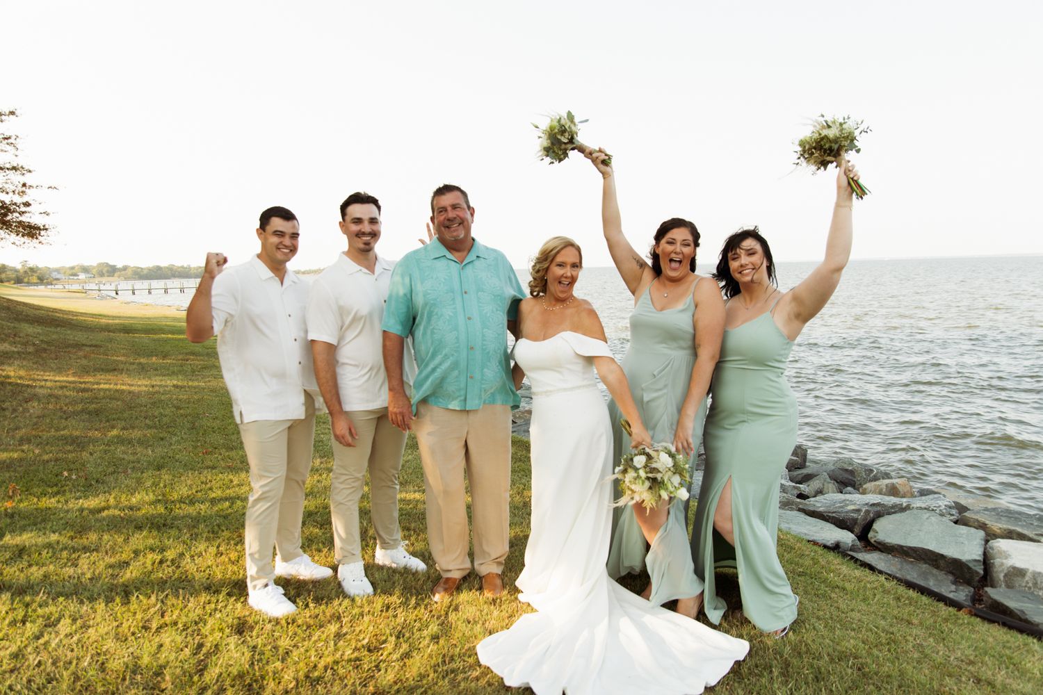 Wedding party posing outdoors by the water, bride in white gown with bridesmaids in sage green dresses.
