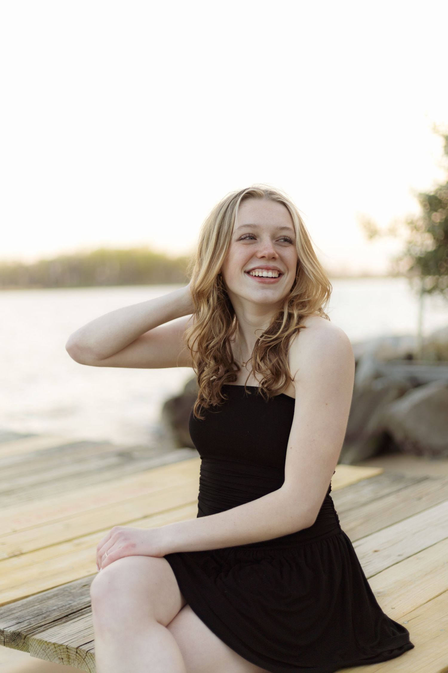 Person in black dress sits on wooden dock at sunset.