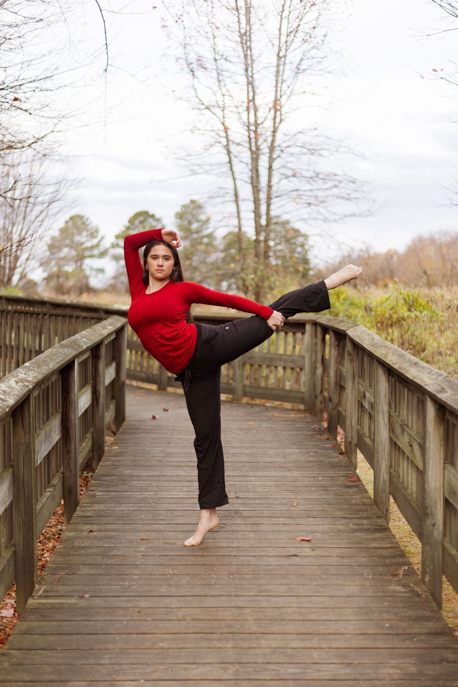 Dancer performs standing split pose on wooden bridge surrounded by winter trees.