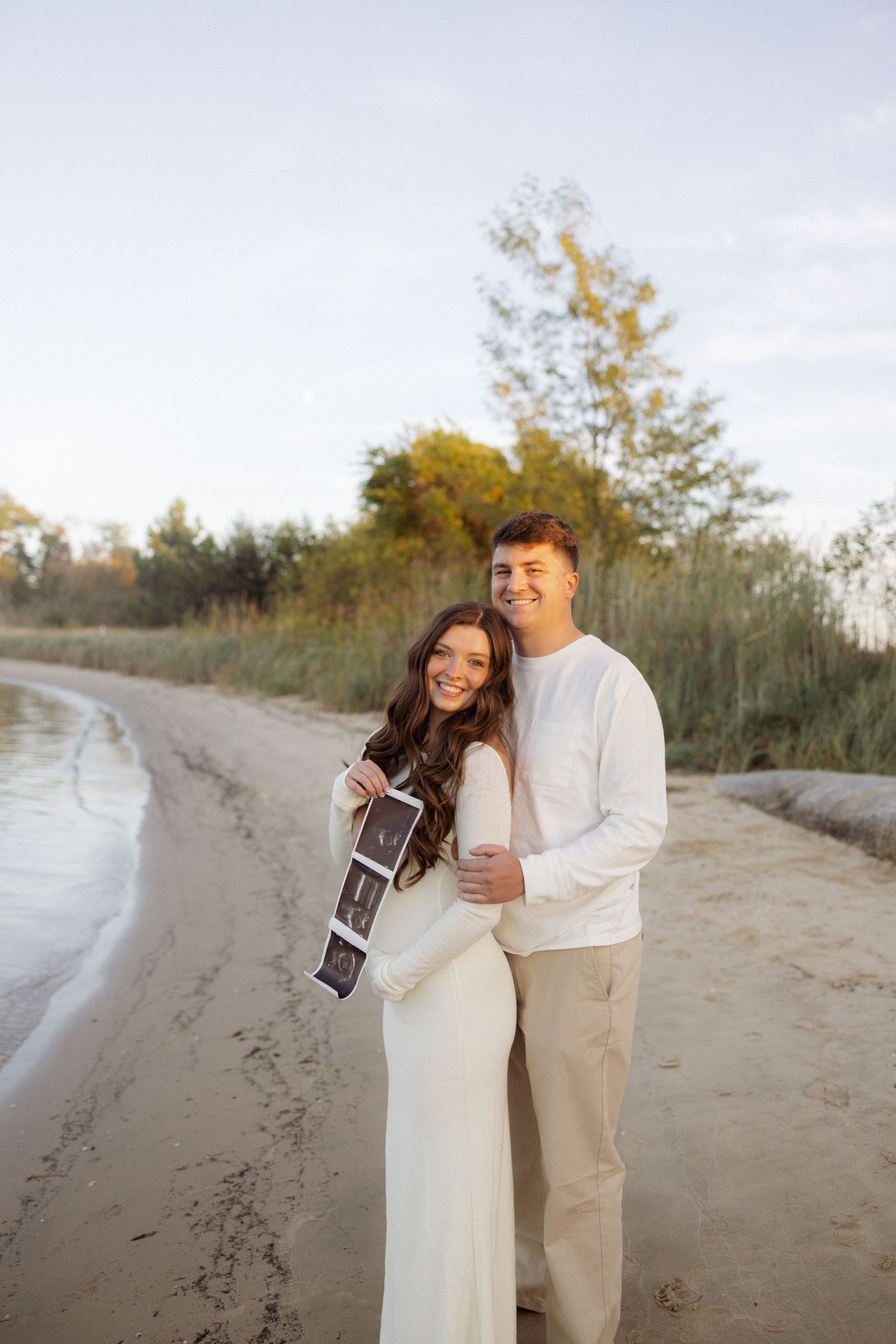 A couple shares a joyful moment on a sandy beach while holding an ultrasound photo.