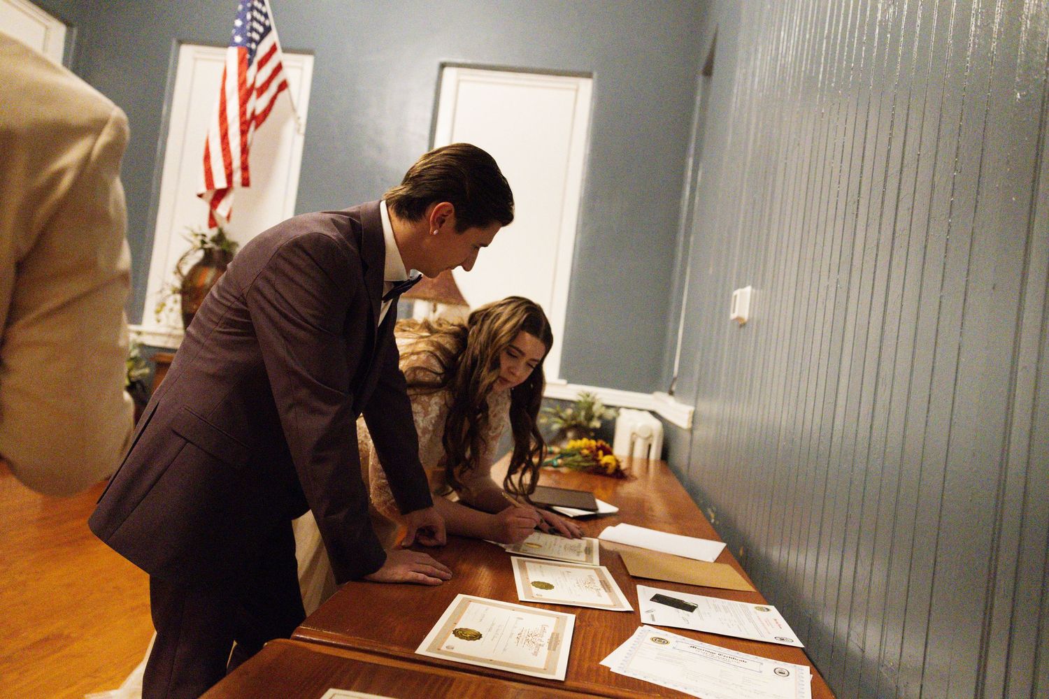 A man and woman in formal attire review documents on a wooden table near an American flag.