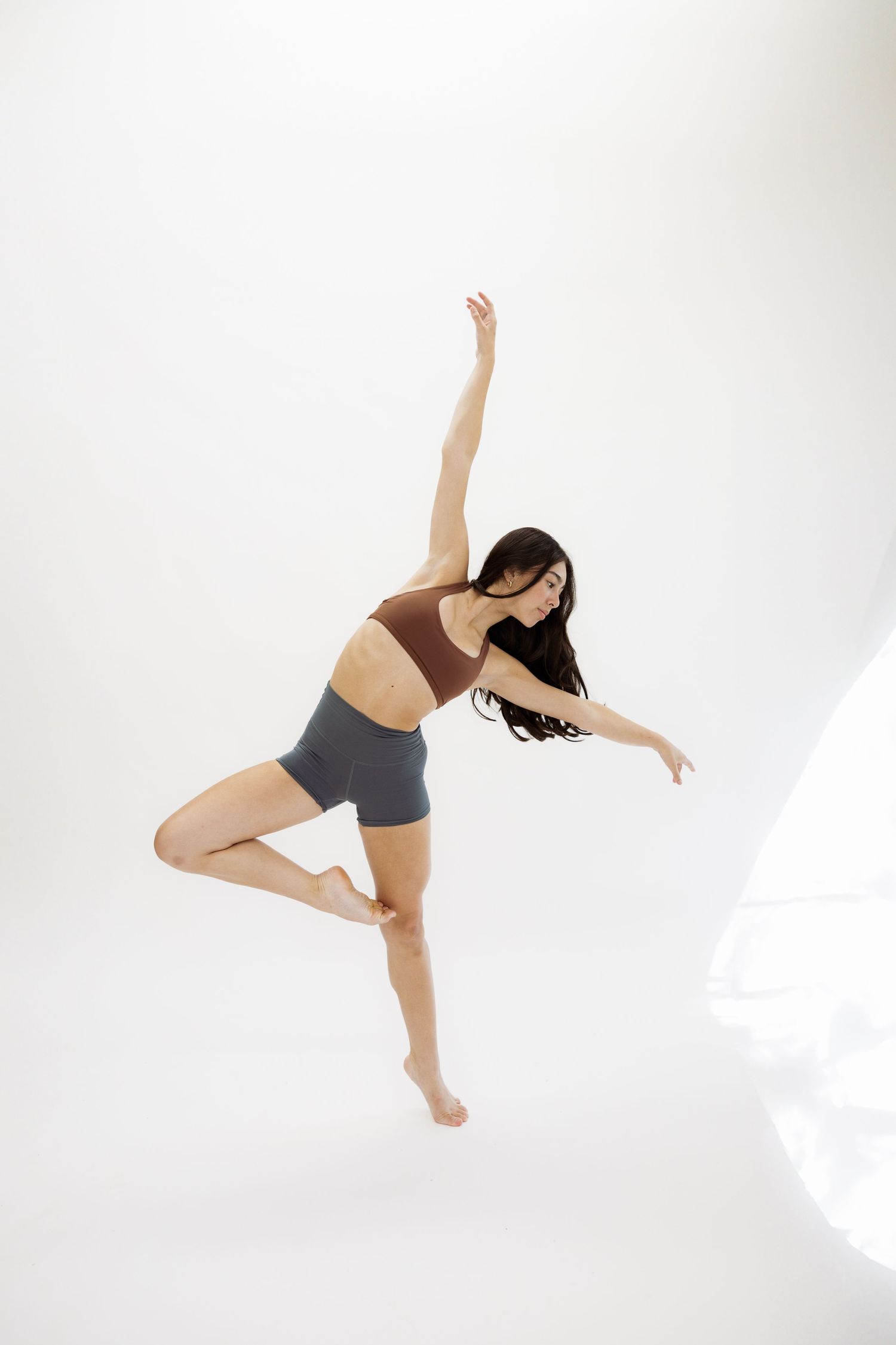 A dancer in brown sports bra and grey shorts performs an elegant pose on a white background.