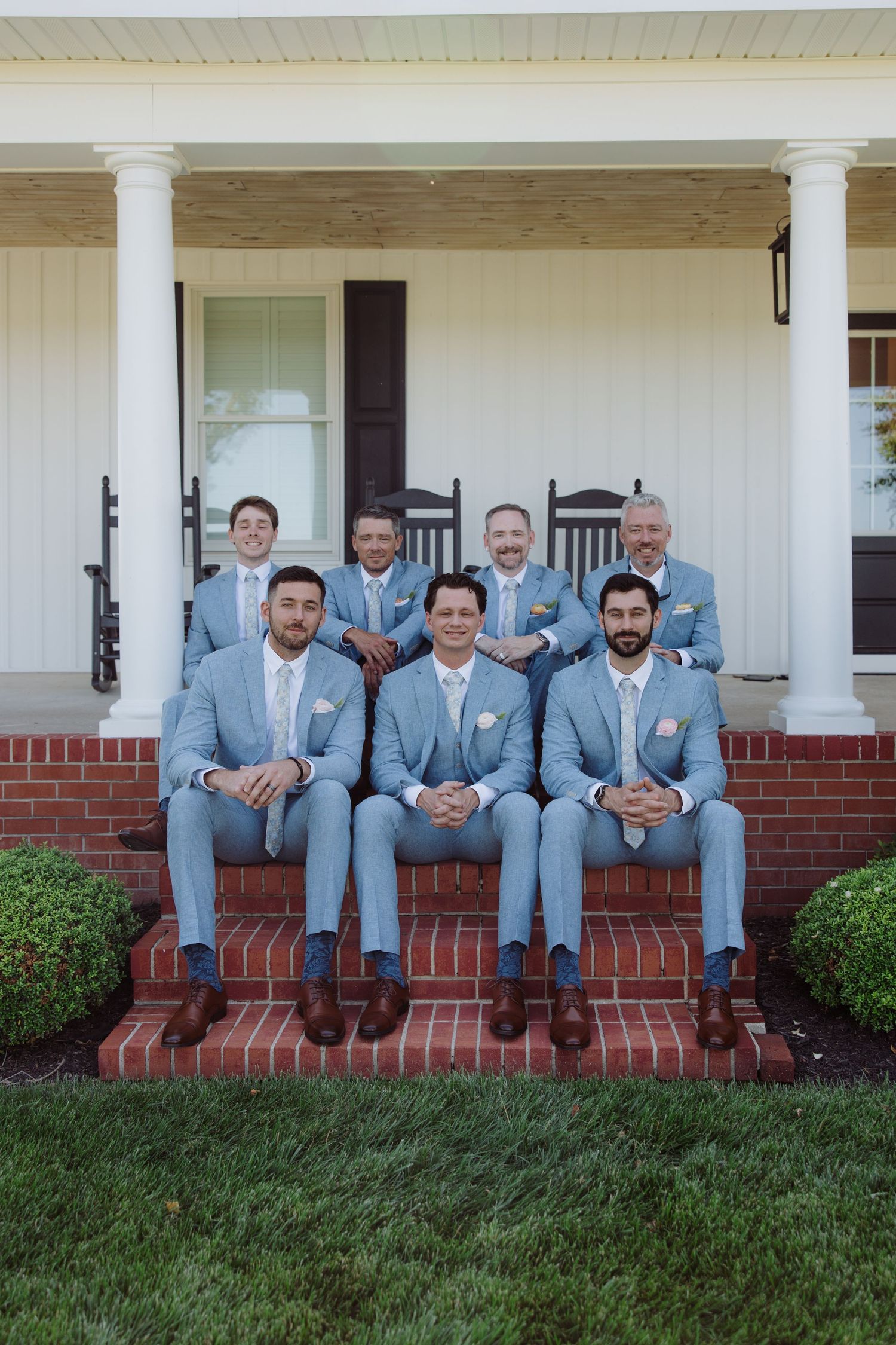Seven groomsmen in light blue suits pose on brick steps of a white farmhouse porch for a wedding photo.
