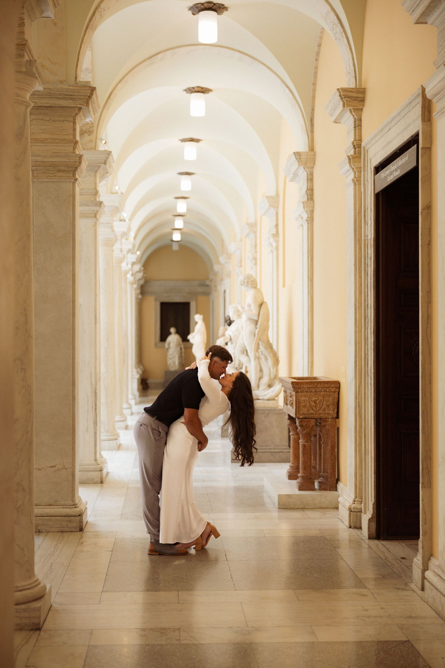 People walk through a bright white arched hallway with ornate columns and wooden doors in a historic building.