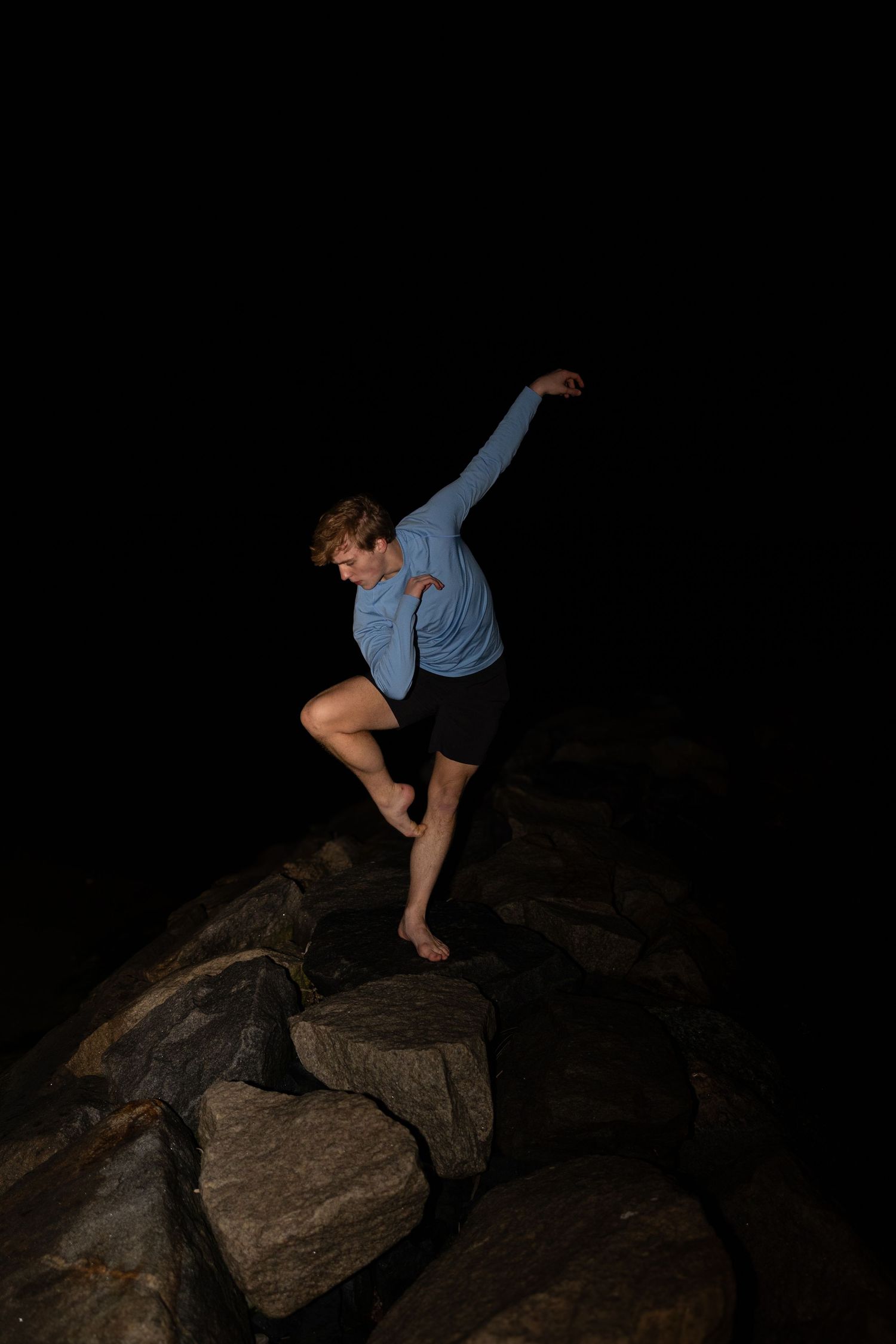 A dancer performs a graceful pose while balancing on rocky terrain at night, wearing blue shirt and black shorts.