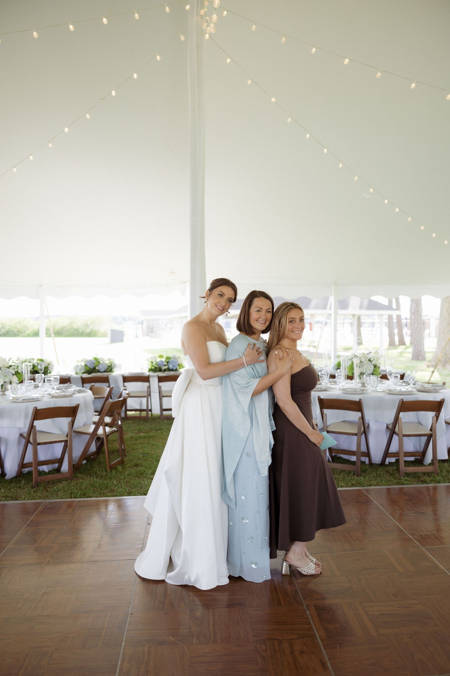 Bride in white gown poses with two women in a decorated wedding tent reception venue.