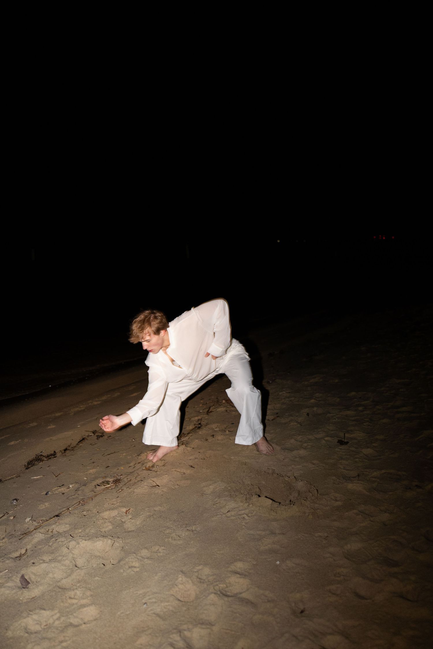 Person in white clothing crouches down on dark sandy ground at night.