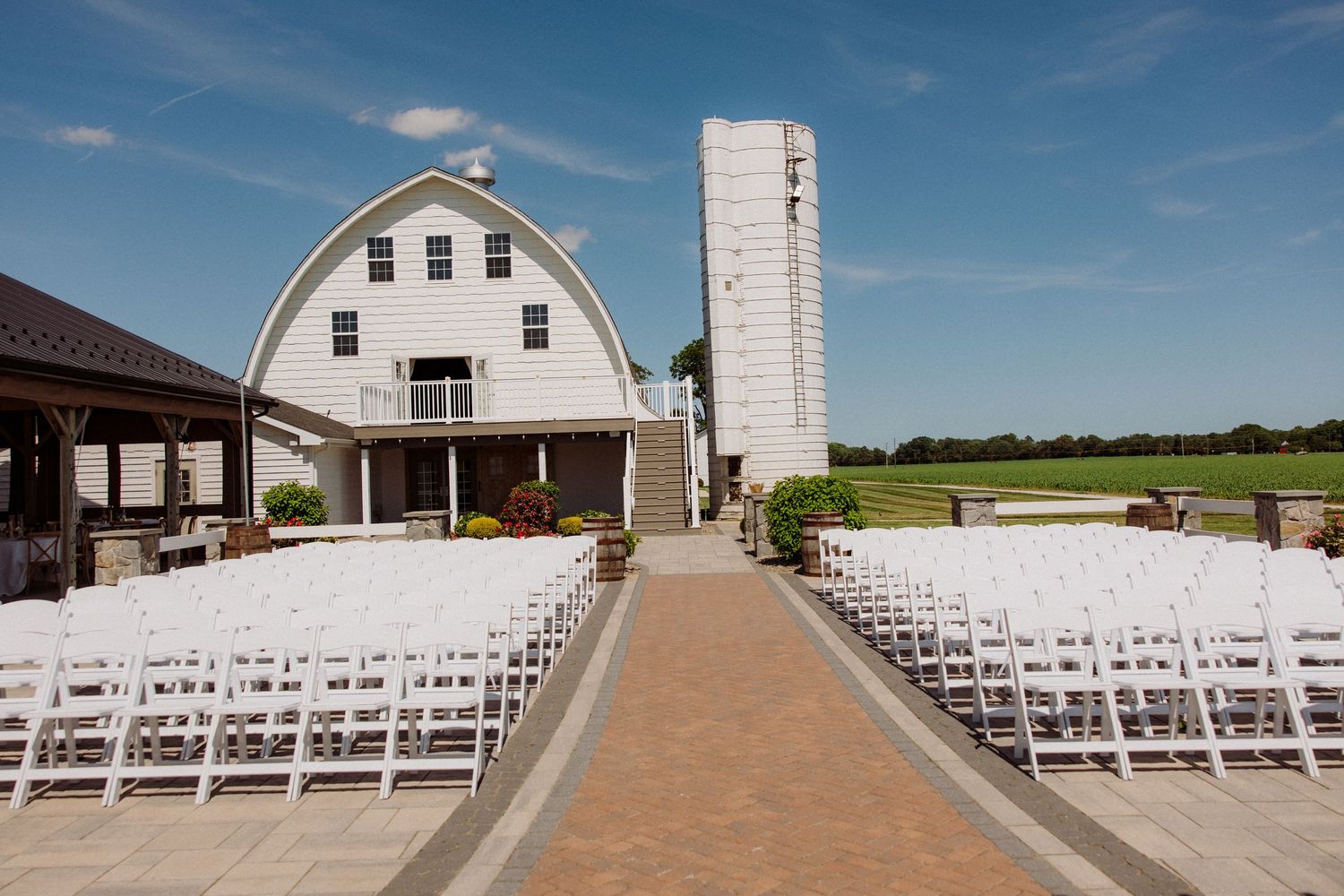 White barn wedding venue with rows of white chairs on brick pathway under blue sky.