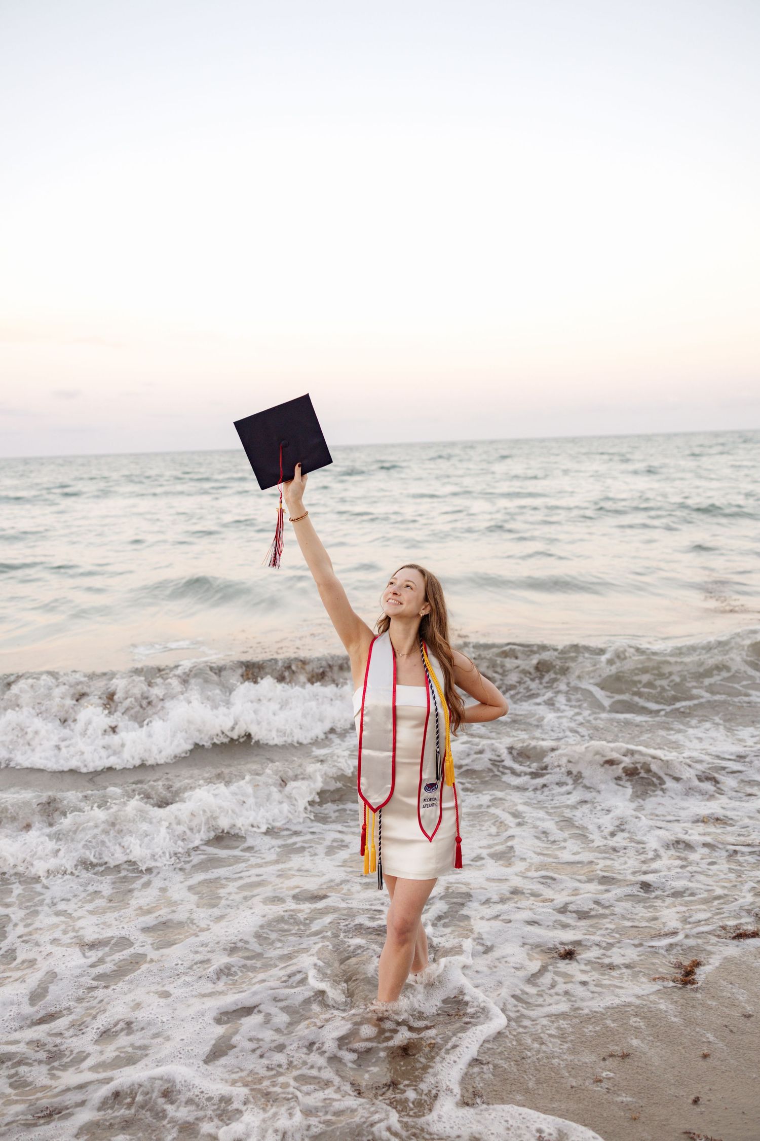 Person in white dress holds graduation cap while standing in ocean waves.