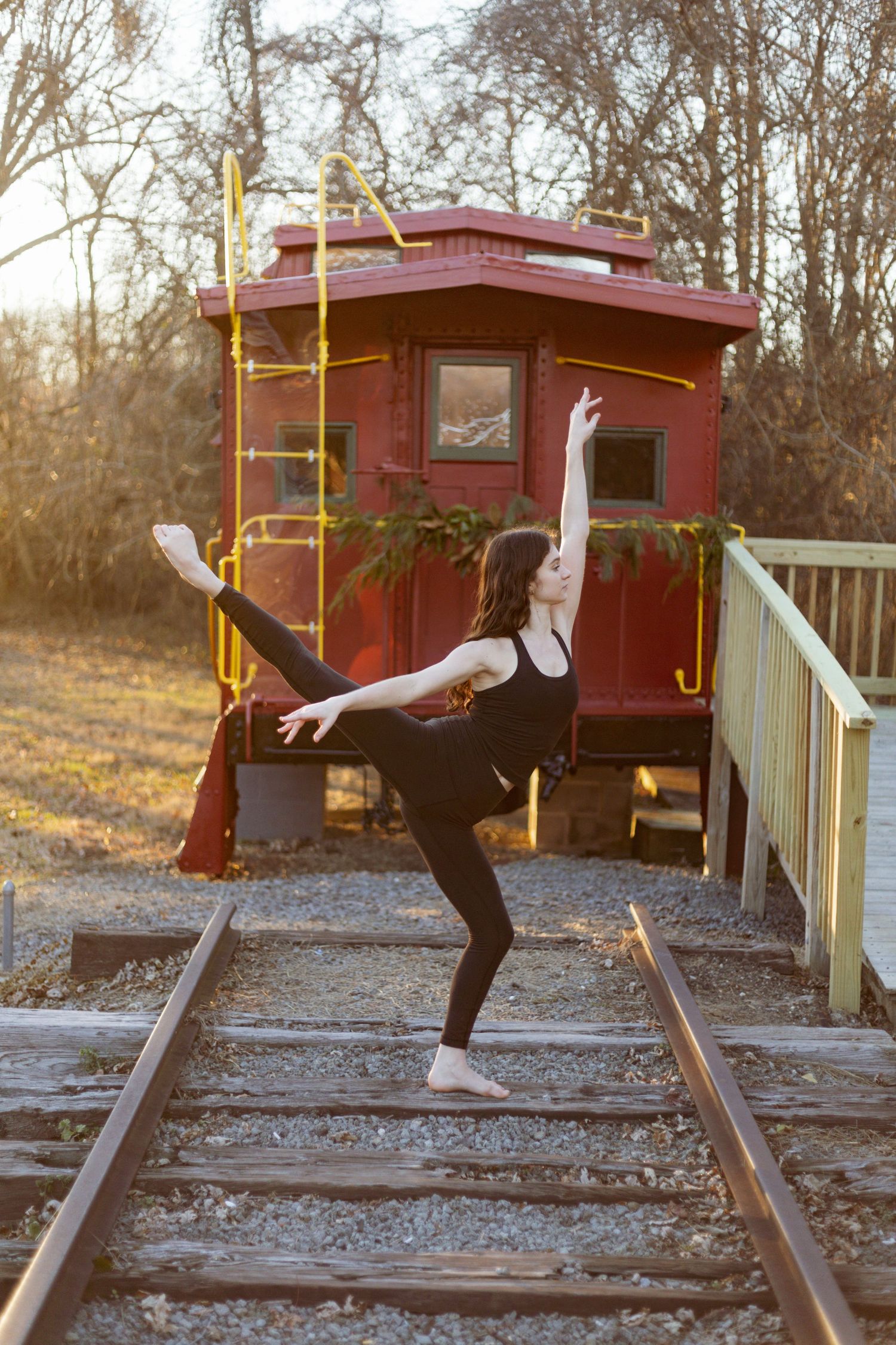 Dancer strikes elegant pose on railroad tracks in front of vintage red caboose at sunset.
