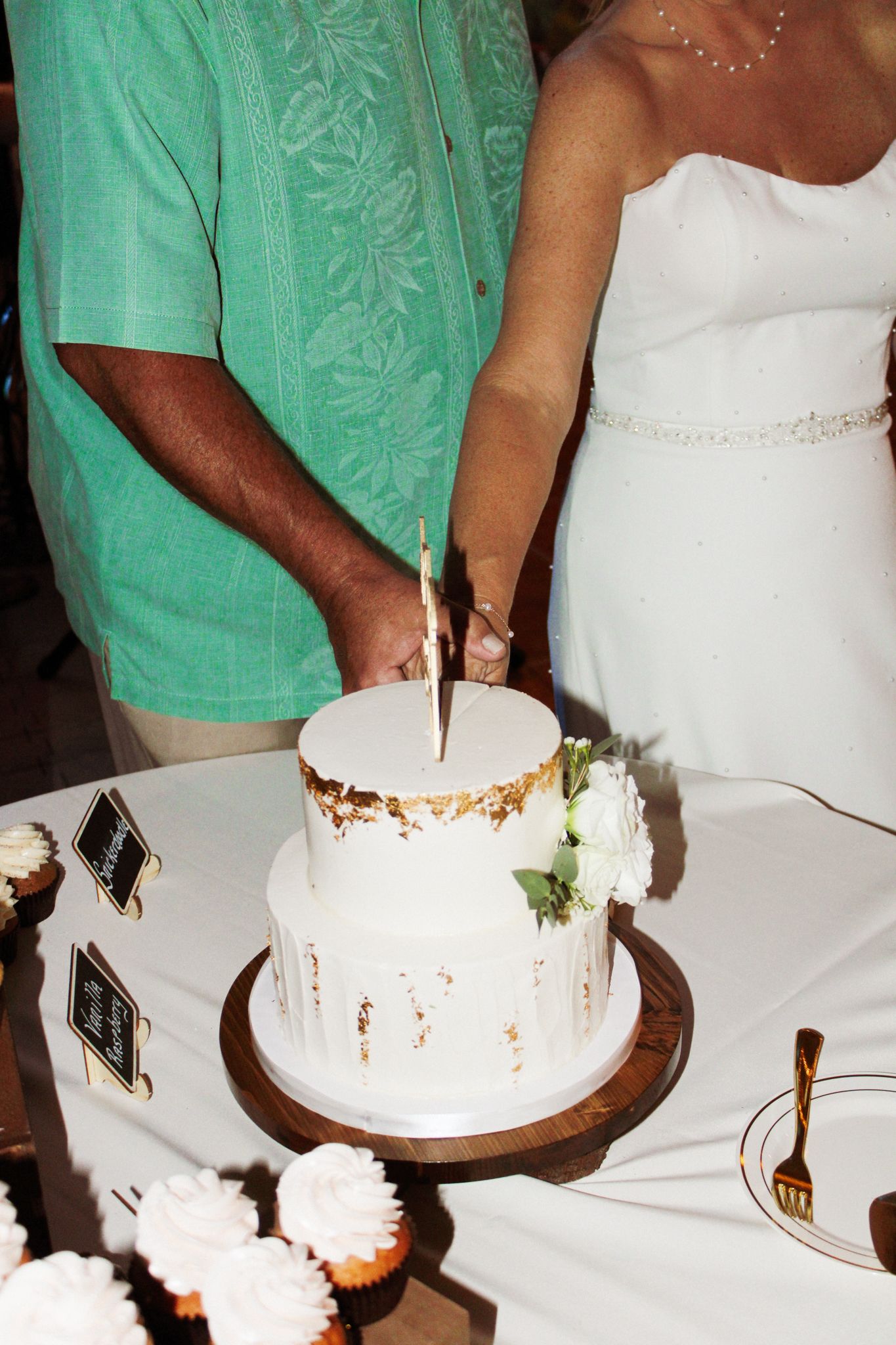 Bride and groom cutting a two-tier white wedding cake with gold leaf accents and white floral decorations.