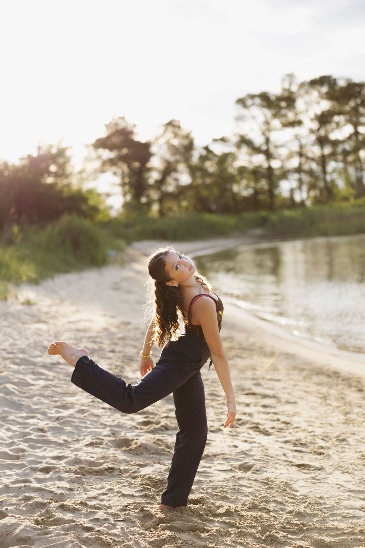 Young woman in black outfit dancing joyfully on a sandy beach at sunset near a tranquil lake and tall trees.