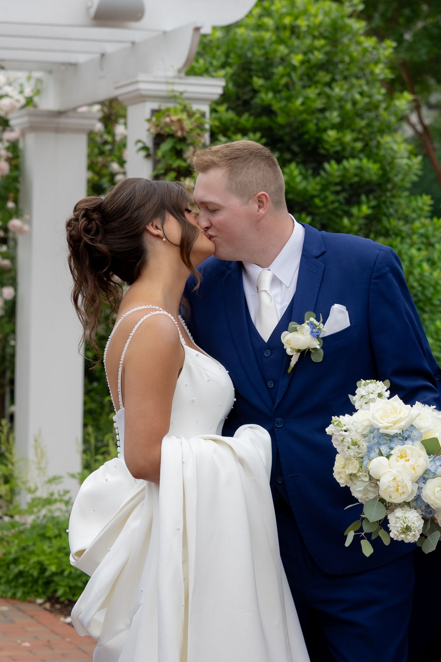 A bride and groom share a romantic kiss outdoors under a white pergola, surrounded by lush greenery at their wedding.