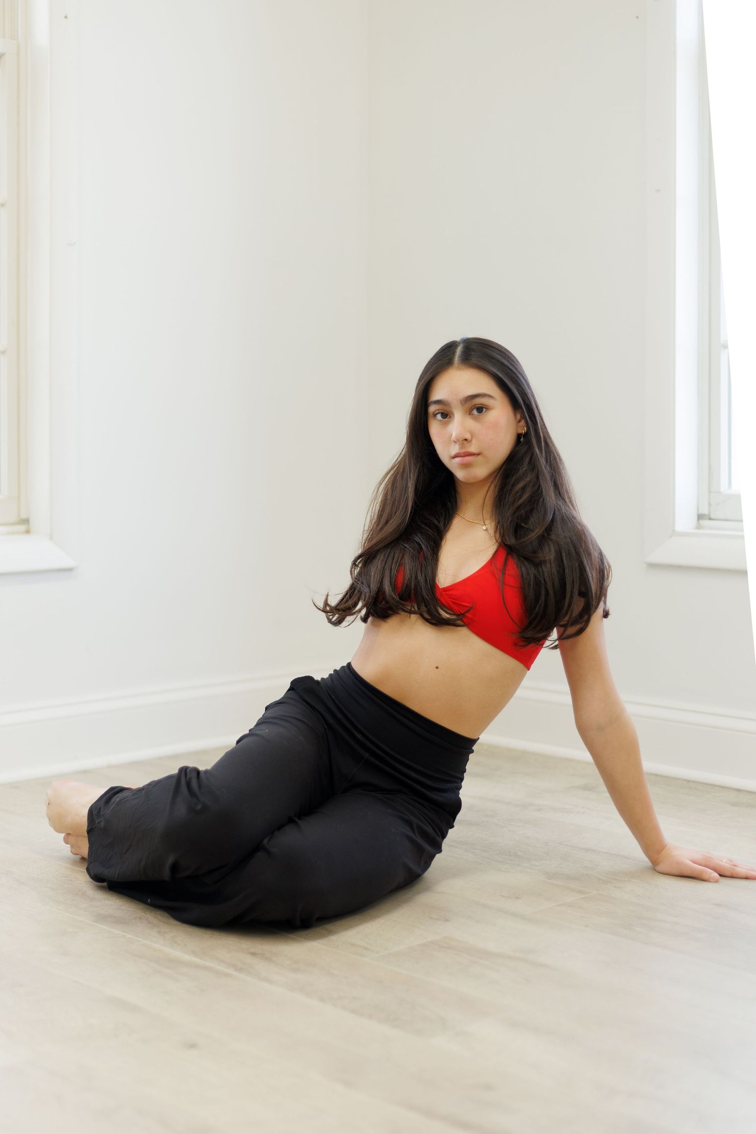 Dancer in red crop top and black pants sits gracefully on light wood floor in bright white studio space.