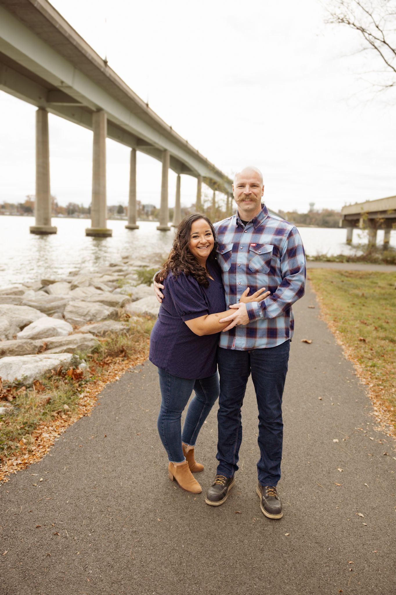 Couple poses together on walking path underneath large bridge structure.