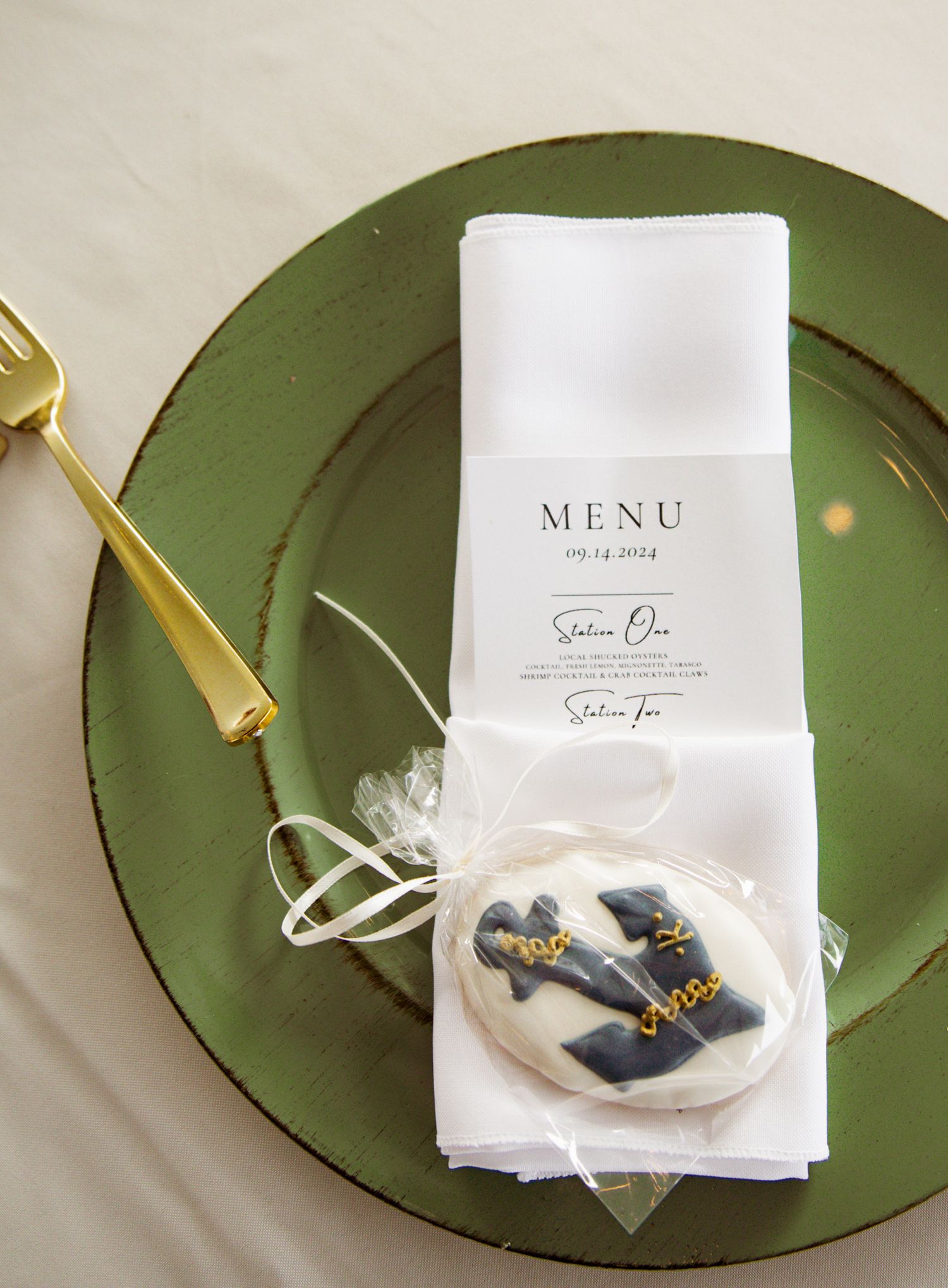 Elegant wedding place setting with green charger plate, gold fork, white menu card, and custom iced sugar cookie favor.