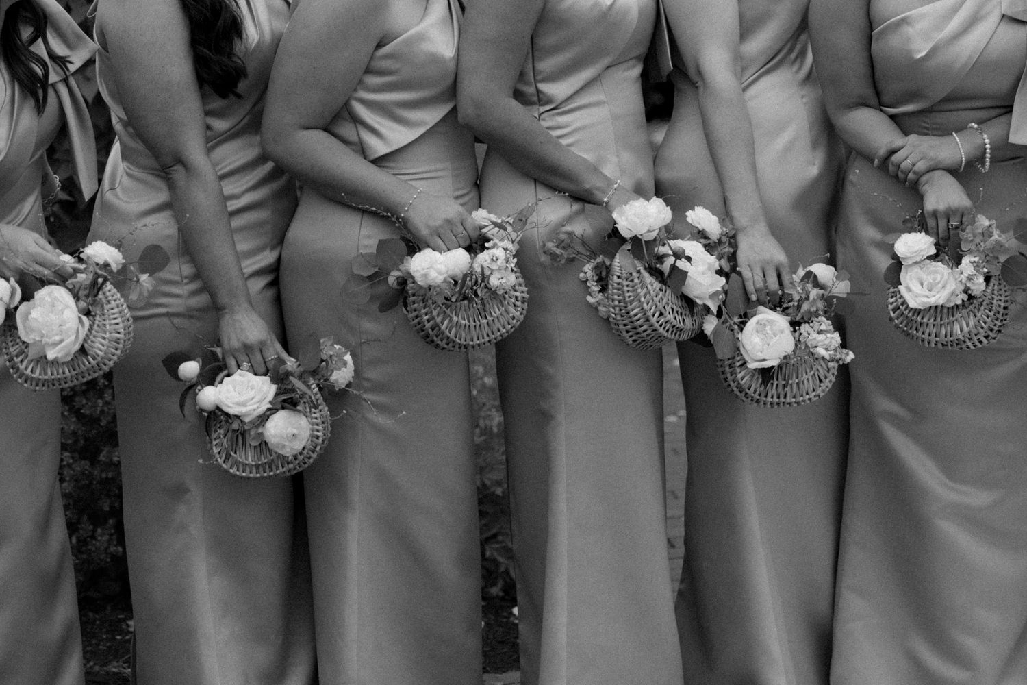Bridesmaids in matching gray gowns holding floral bouquets with woven accents, captured in elegant black and white photography.