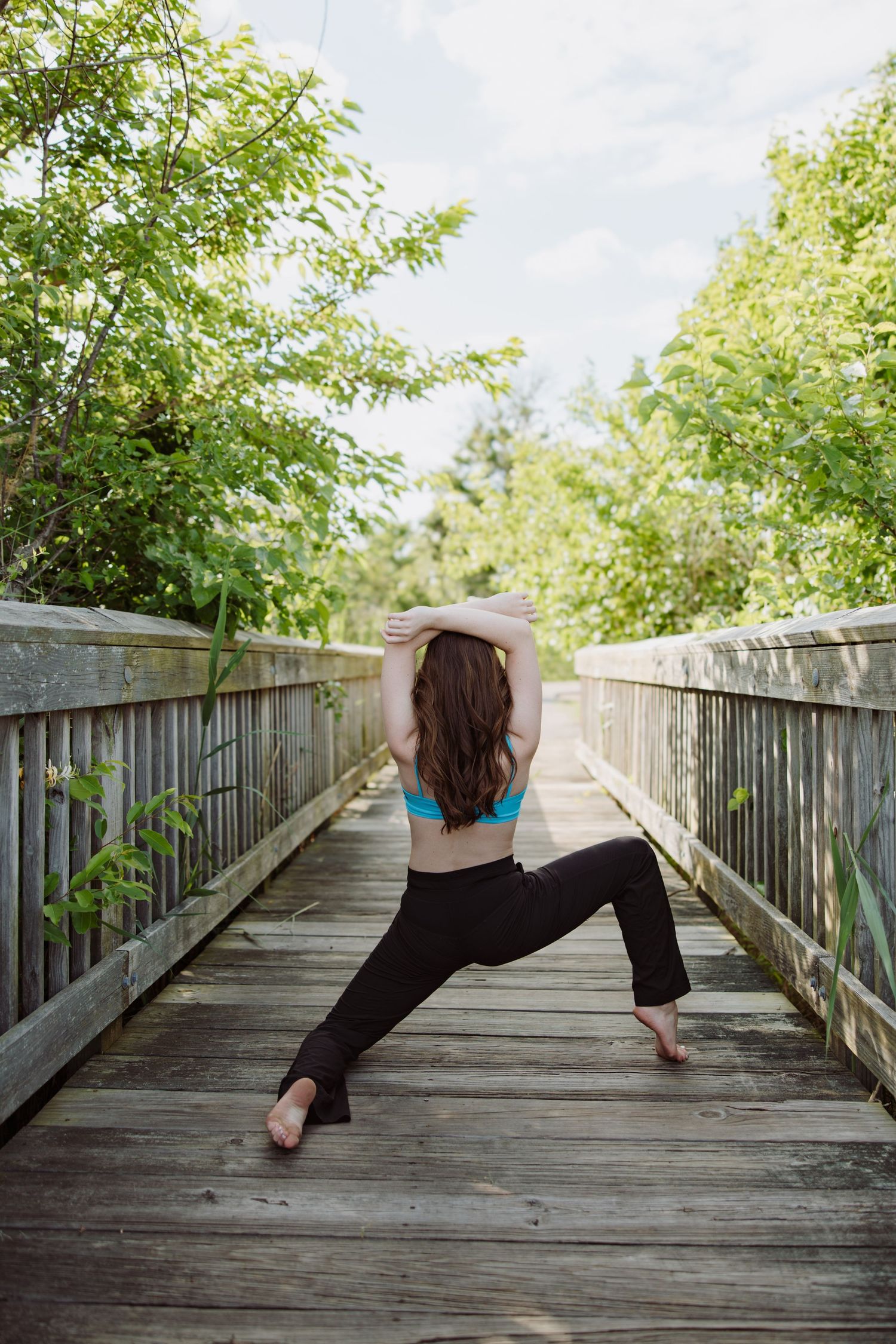 Dancer in turquoise top and black pants strikes a pose on wooden boardwalk surrounded by green foliage.