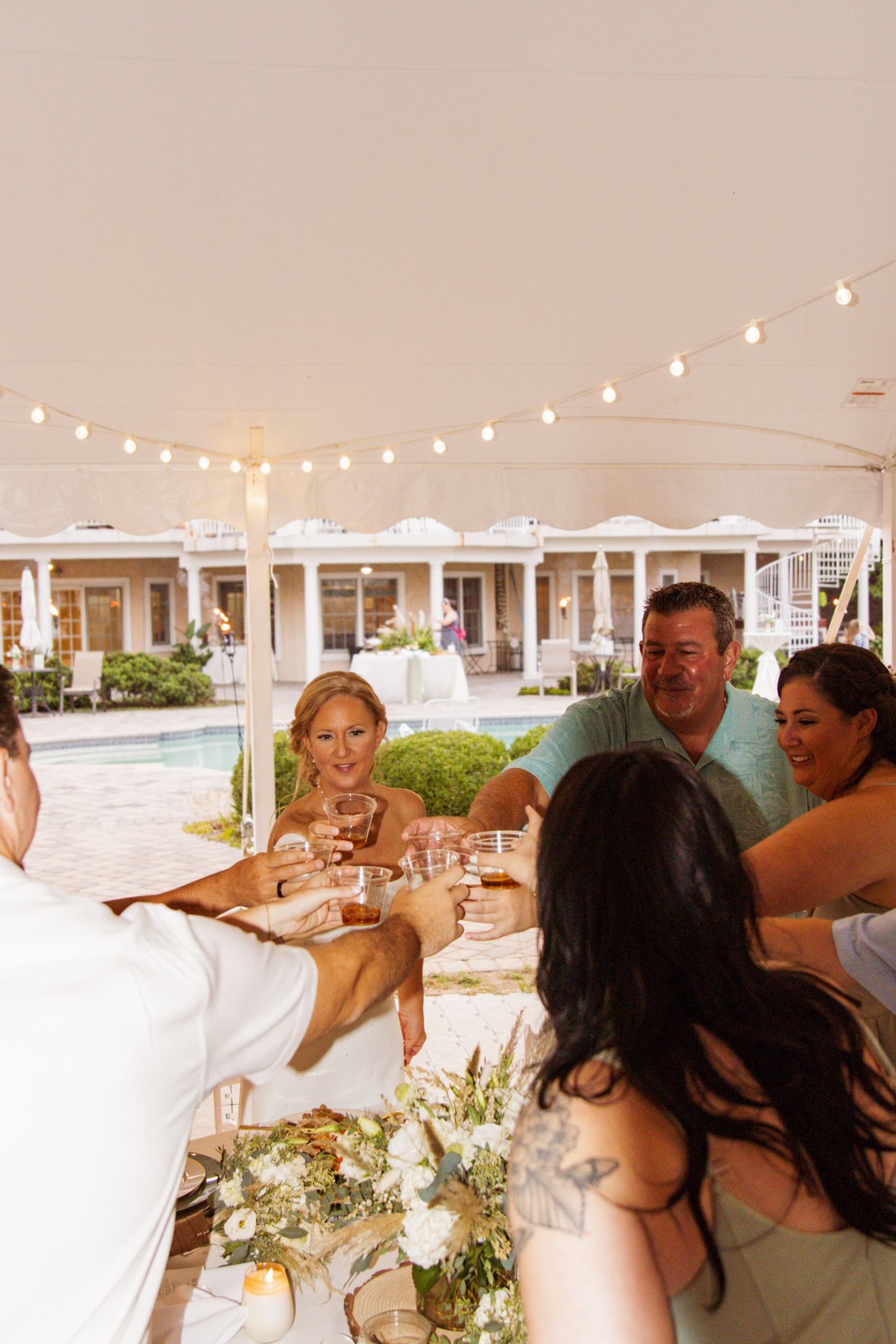 Wedding guests raising glasses in a toast under a string-lit tent by a pool at an outdoor reception.