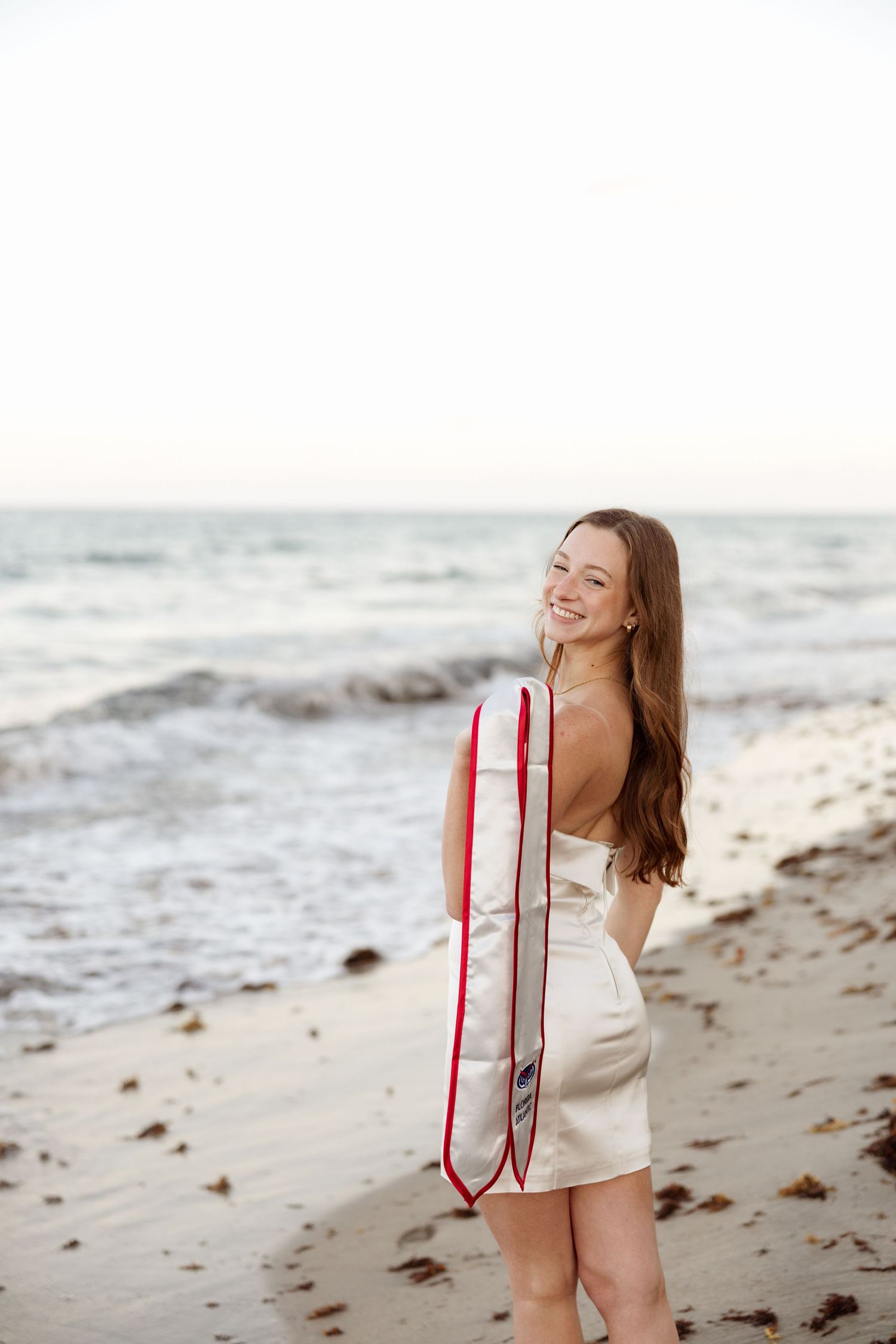 Person in white dress holds surfboard on beach at sunset.