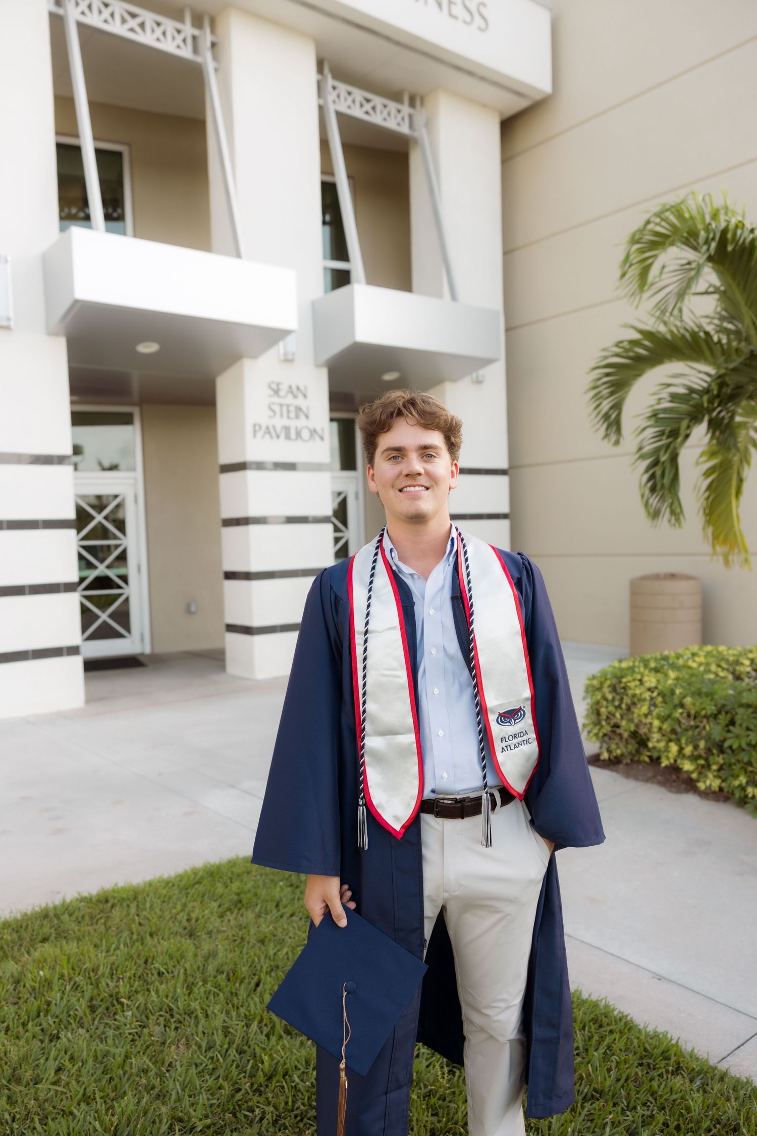 Graduate in navy blue cap and gown with decorative stole standing outside building entrance.