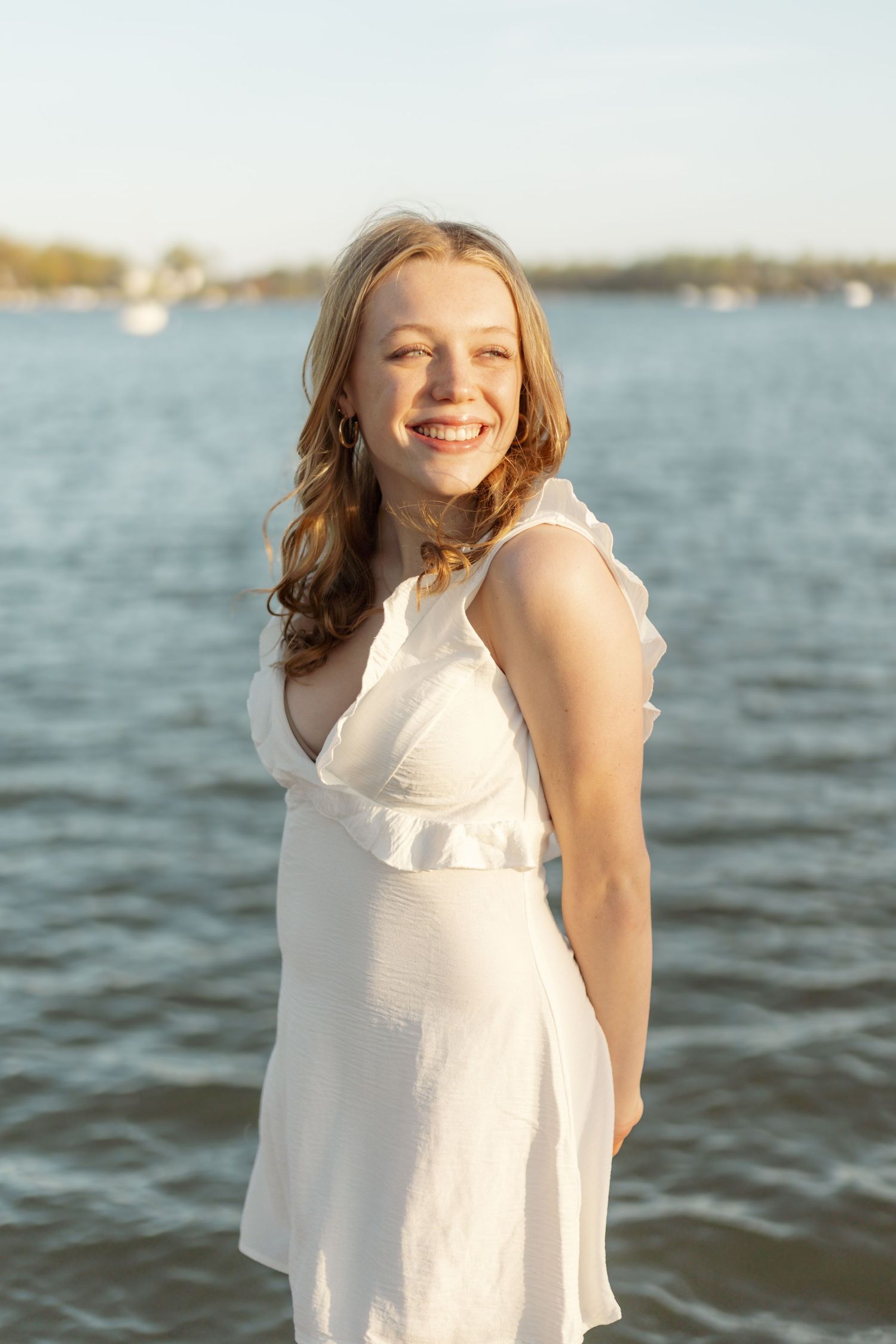Person wearing white dress smiles by water during golden hour.