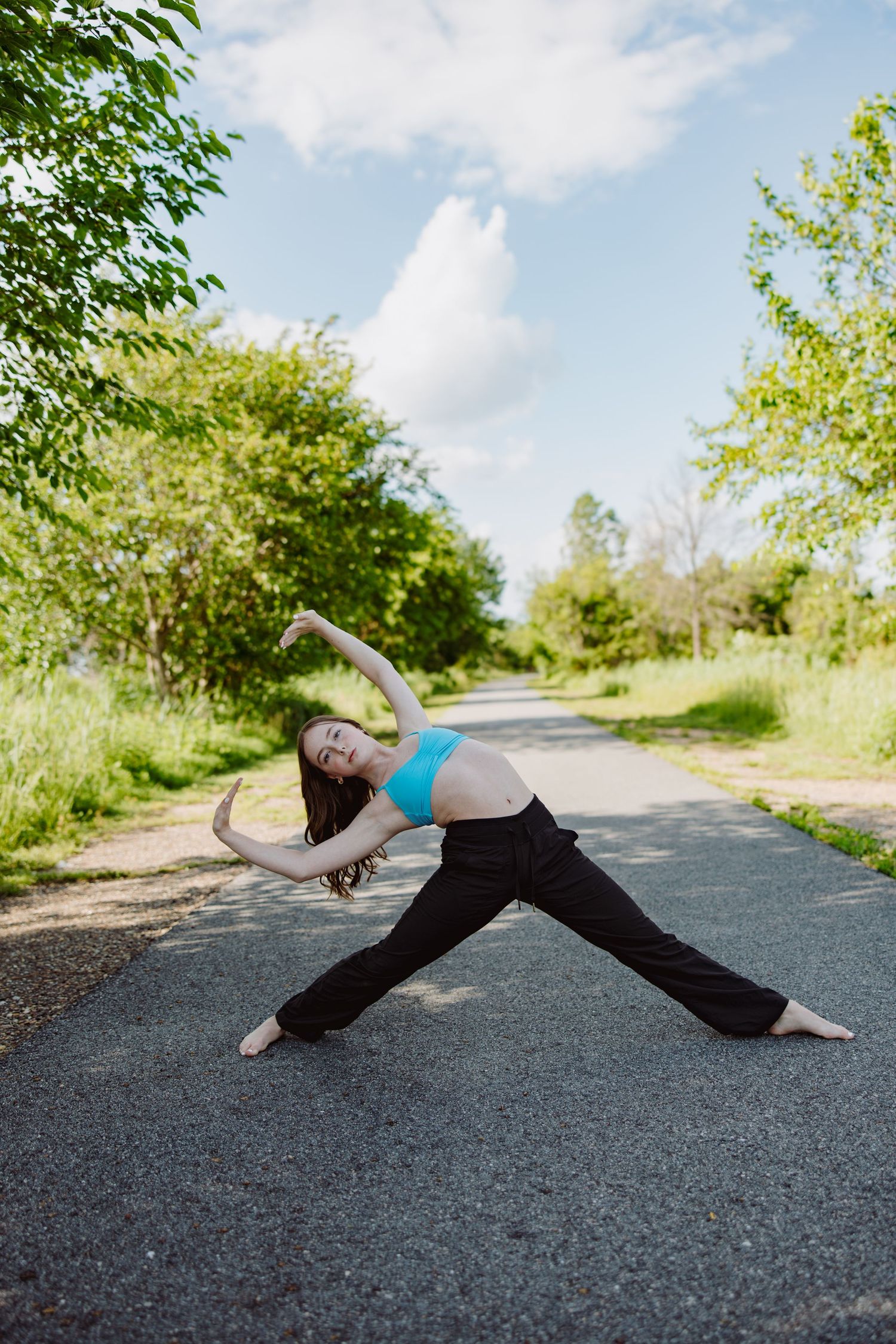 A dancer in a blue top and black pants performs a graceful pose on a tree-lined path on a sunny day.