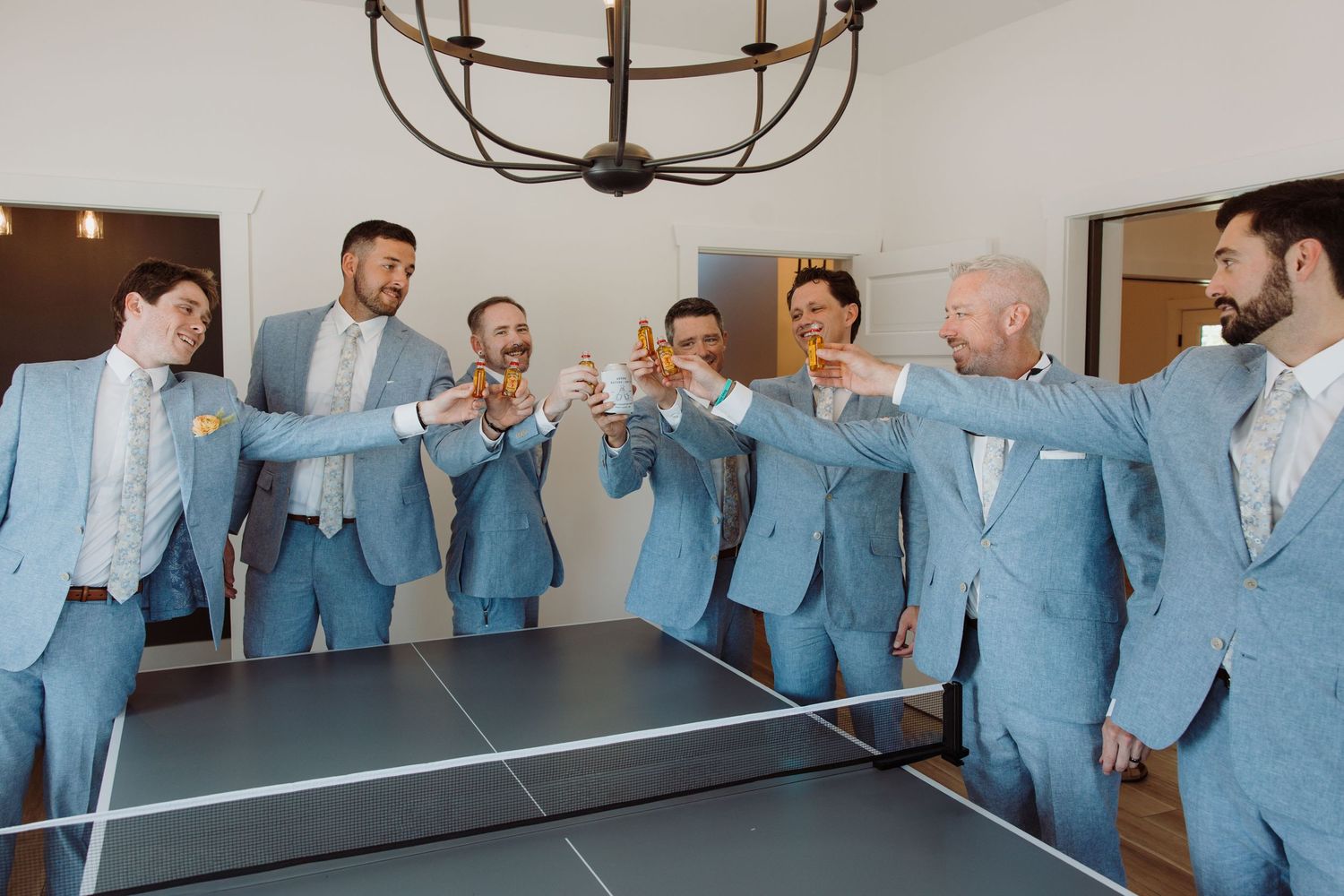 Groomsmen in light blue suits toasting with champagne glasses around a ping pong table before a wedding ceremony.