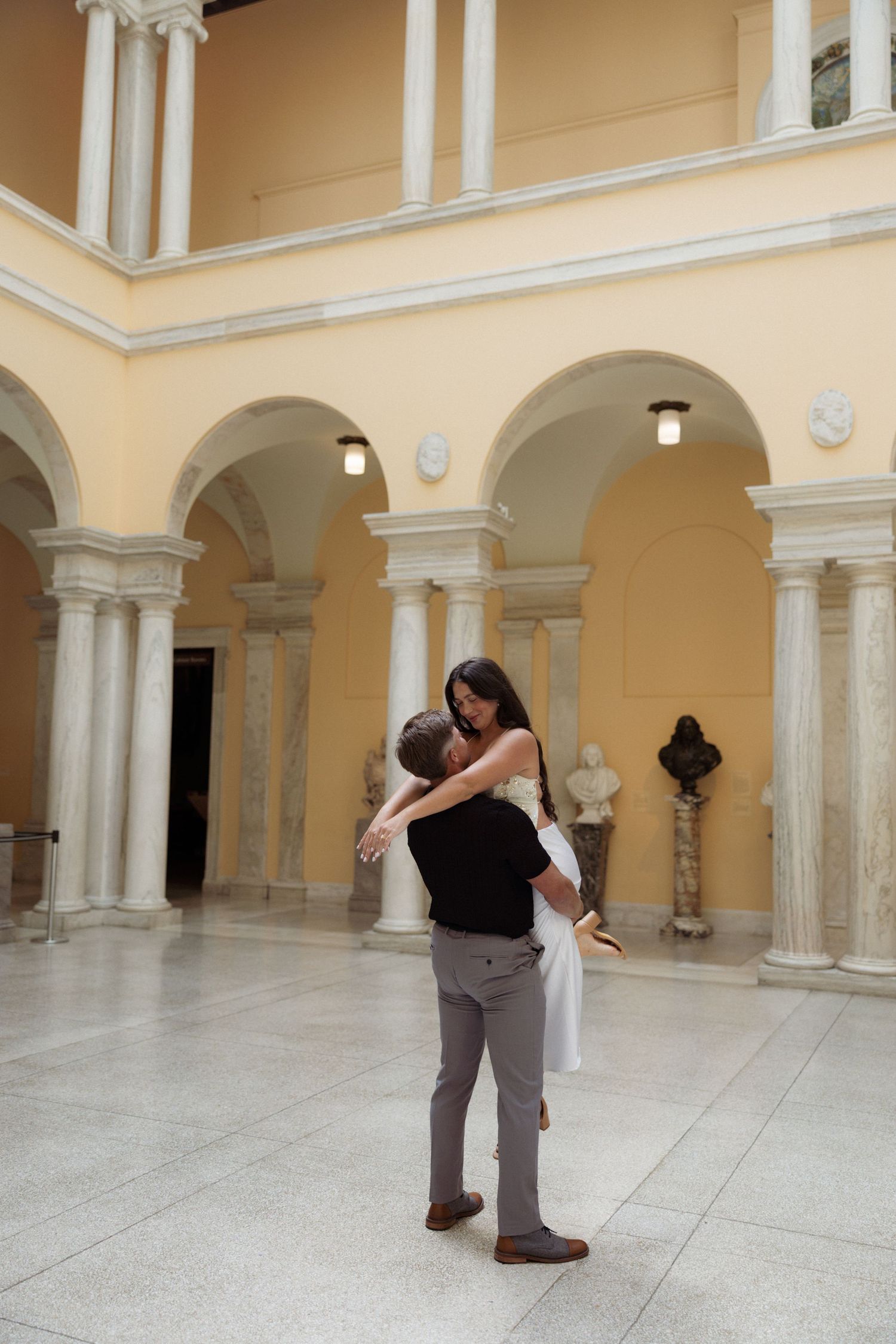 Couple embracing in elegant neoclassical building with arched colonnade, white marble columns, and warm yellow walls.