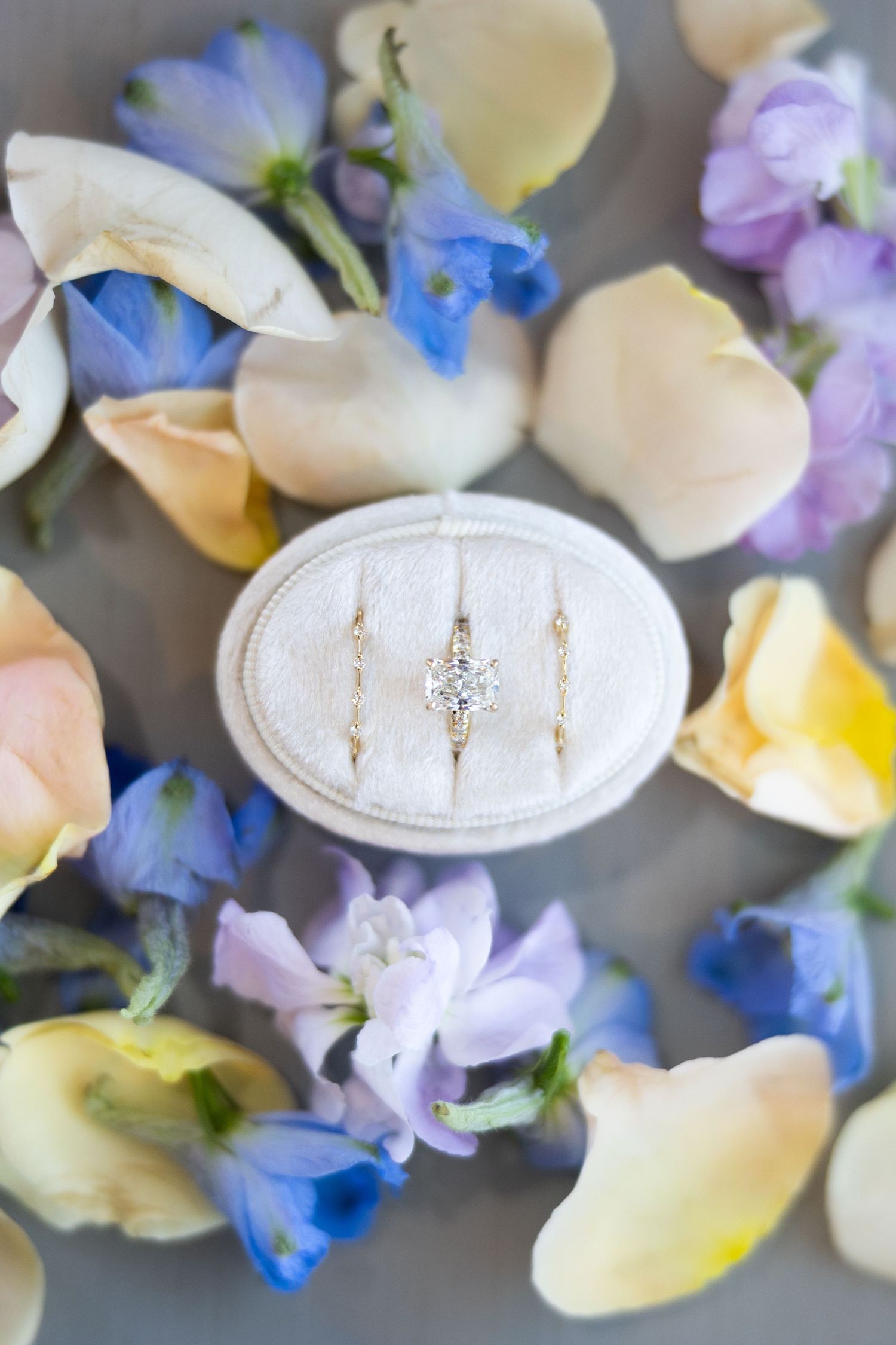 Gold engagement ring with diamond solitaire displayed in velvet box surrounded by colorful spring flower petals.