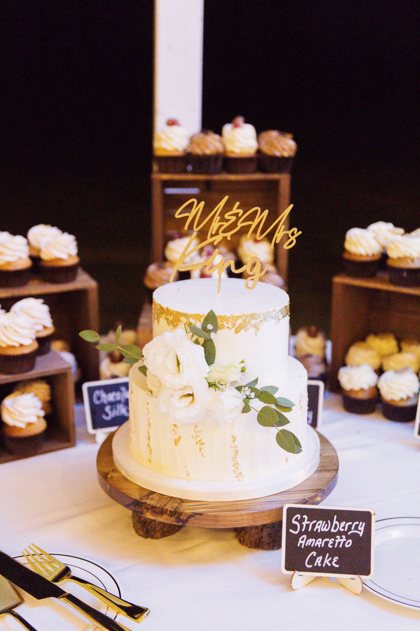 Two-tier white and gold wedding cake with floral decor, surrounded by cupcakes, labeled Strawberry Amaretto Cake.